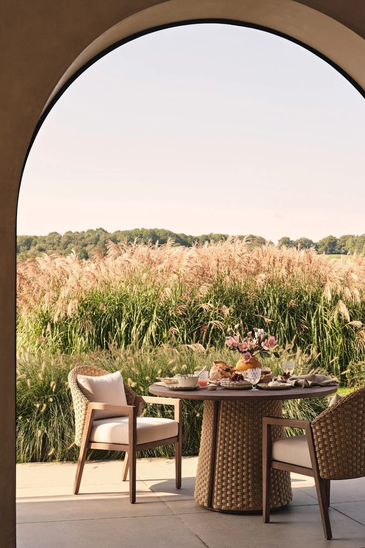 Round outdoor dining table with woven rattan base and matching chairs styled with flowers and tableware beneath an arched terrace.