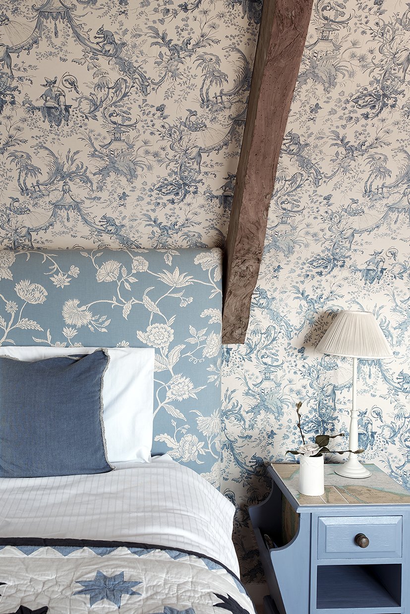 A bed with a patterned floral blue and white headboard with patterned wallpaper behind and an old wooden beam