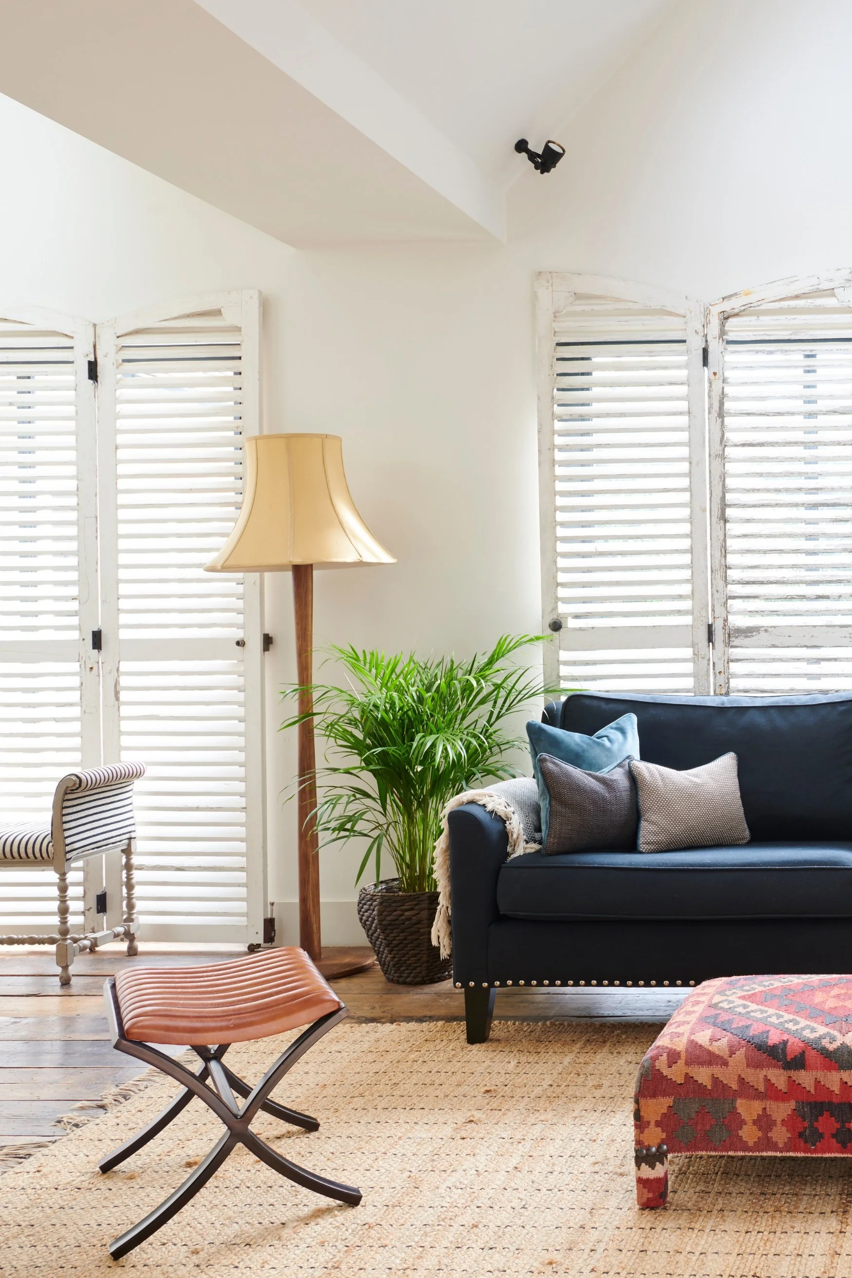 Living room with a blue sofa red ottoman a potted plant and wooden lamp with linen shade. There is a leather stool on a jute rug.