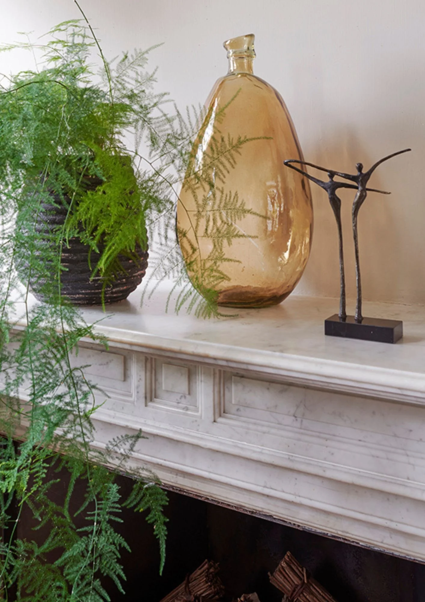 Stylish home decor vase in amber glass, an asparagus fern in a black ribbed pot and two dancing figurines on a marble mantle.