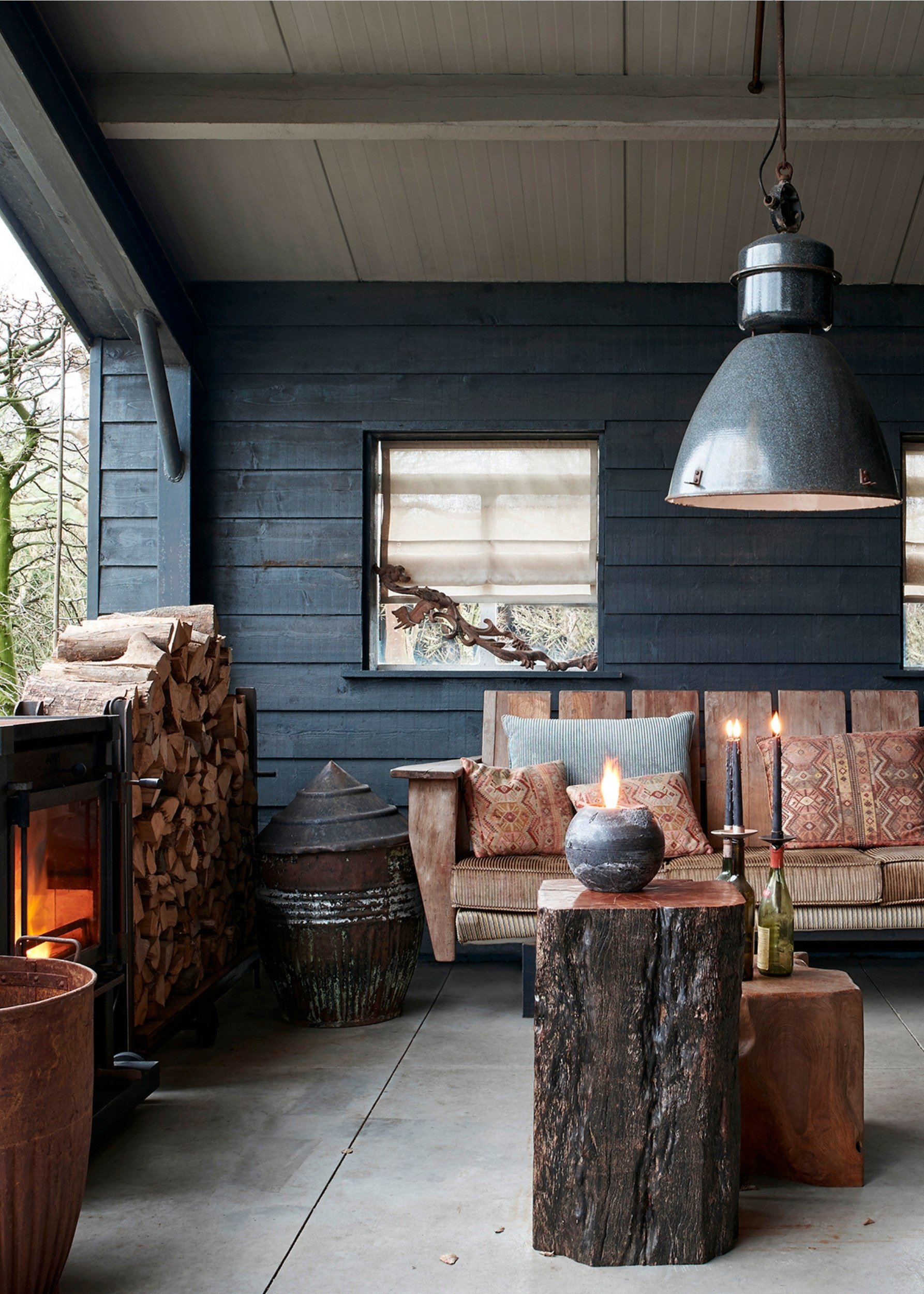 An outdoor undercover area with a woodturning stove next to a pile of logs. There is an outdoor wooden sofa and two side tables made from tree trunks