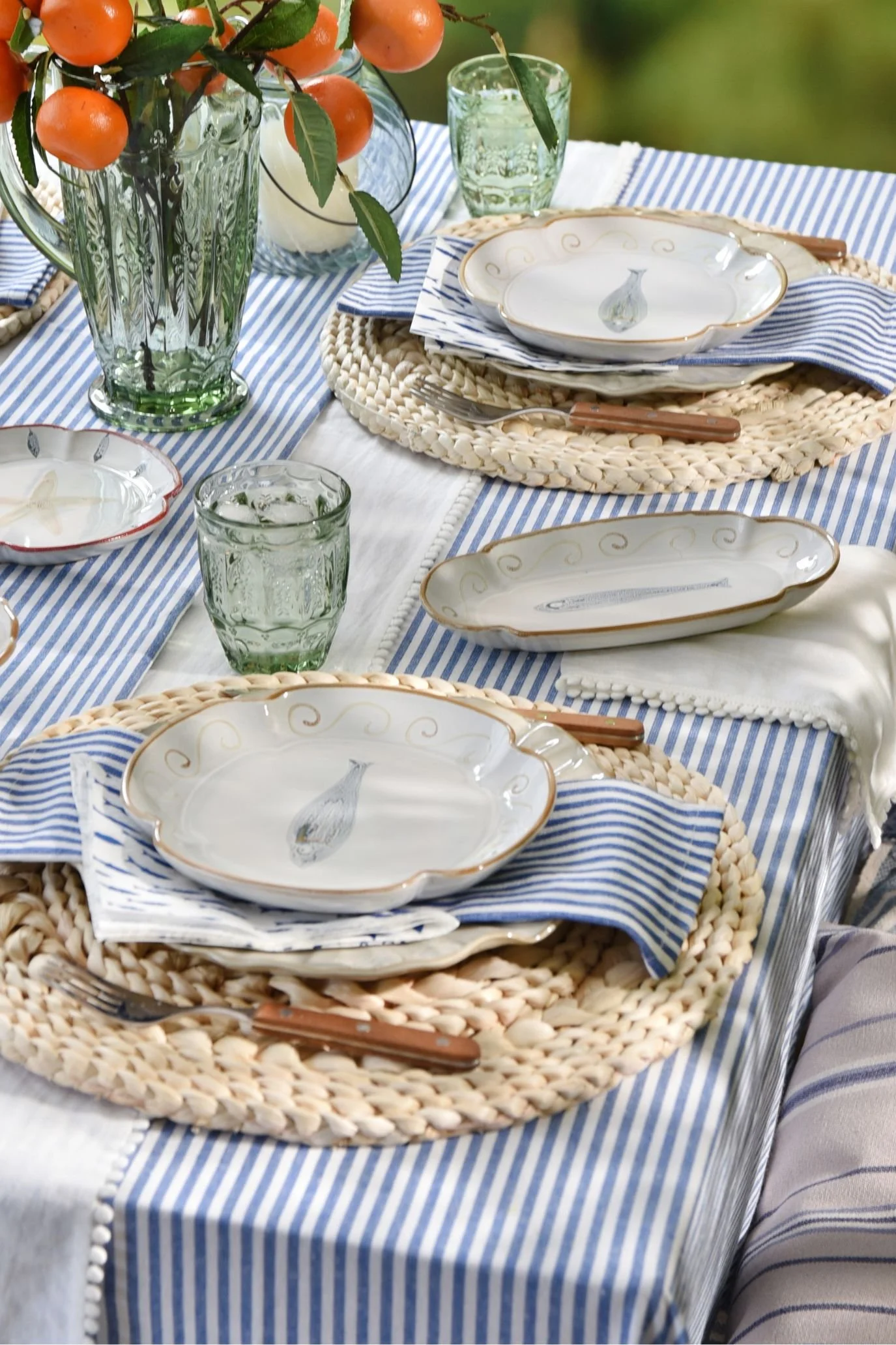 Layered summer table with stoneware plates, woven placemats, green glassware and wooden-handled cutlery.