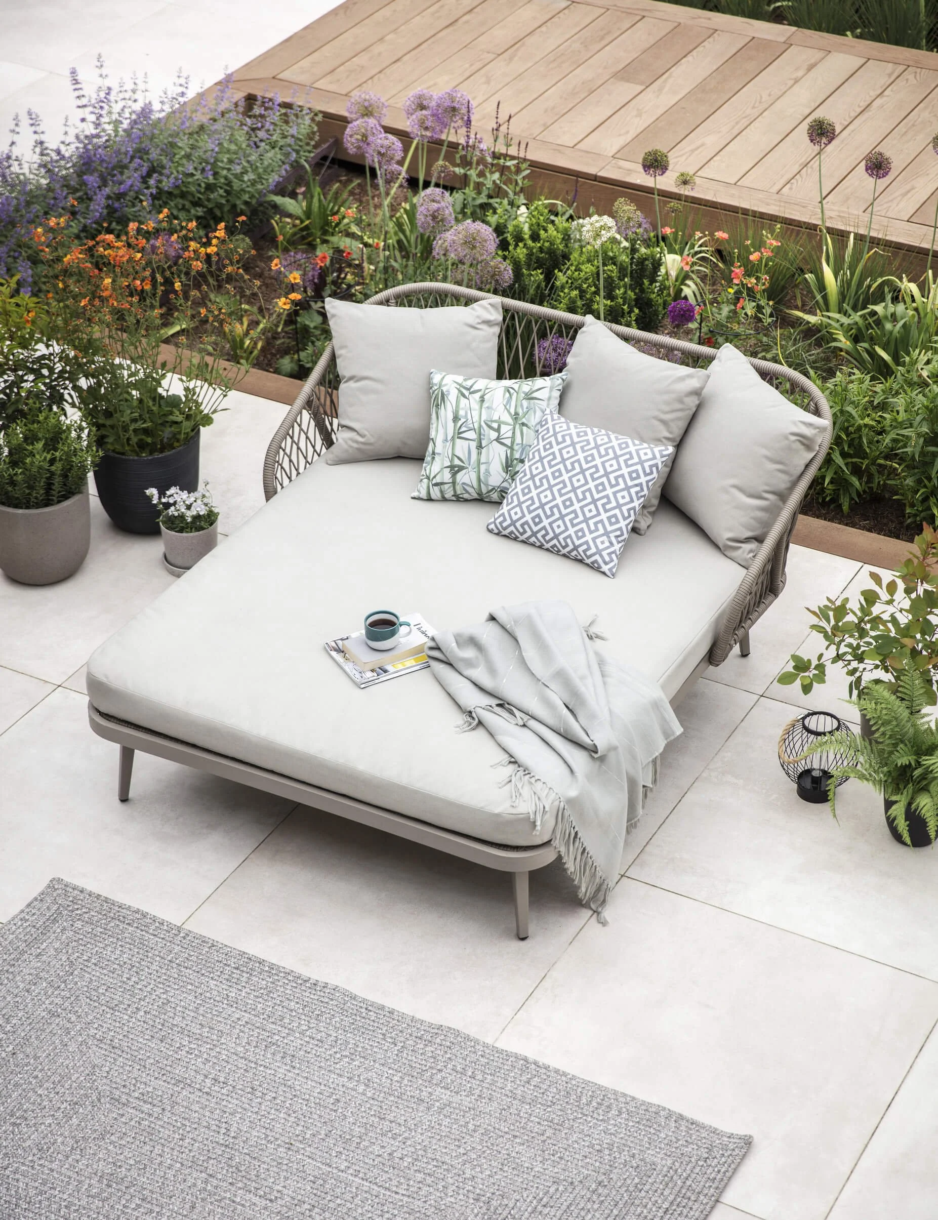 outdoor day bed with grey rope style sides and five cushions on a terrace with pots and a planted raised bed with flowers