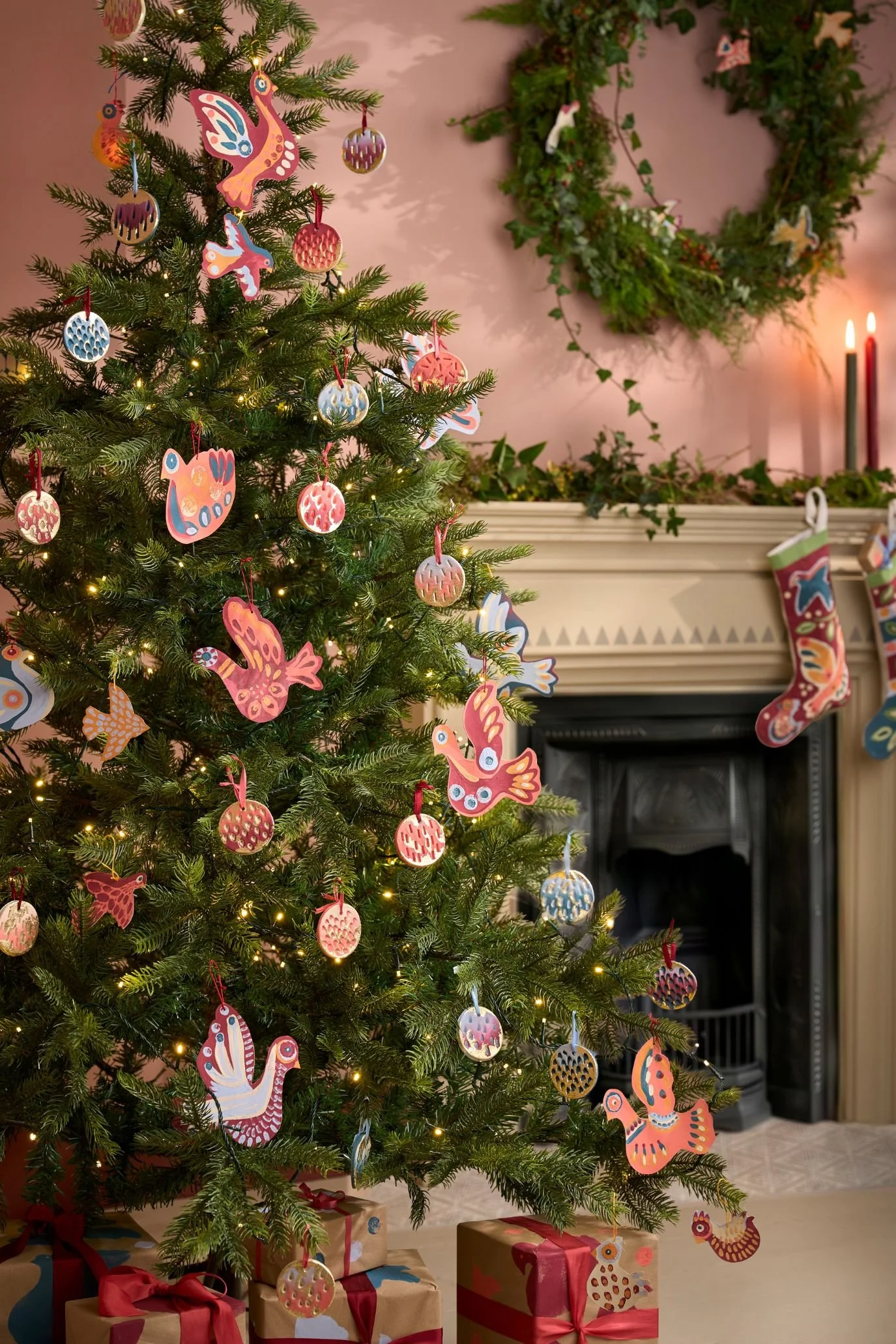 Christmas tree decorated with colourful painted birds and patterned baubles beside a fireplace and greenery wreath.