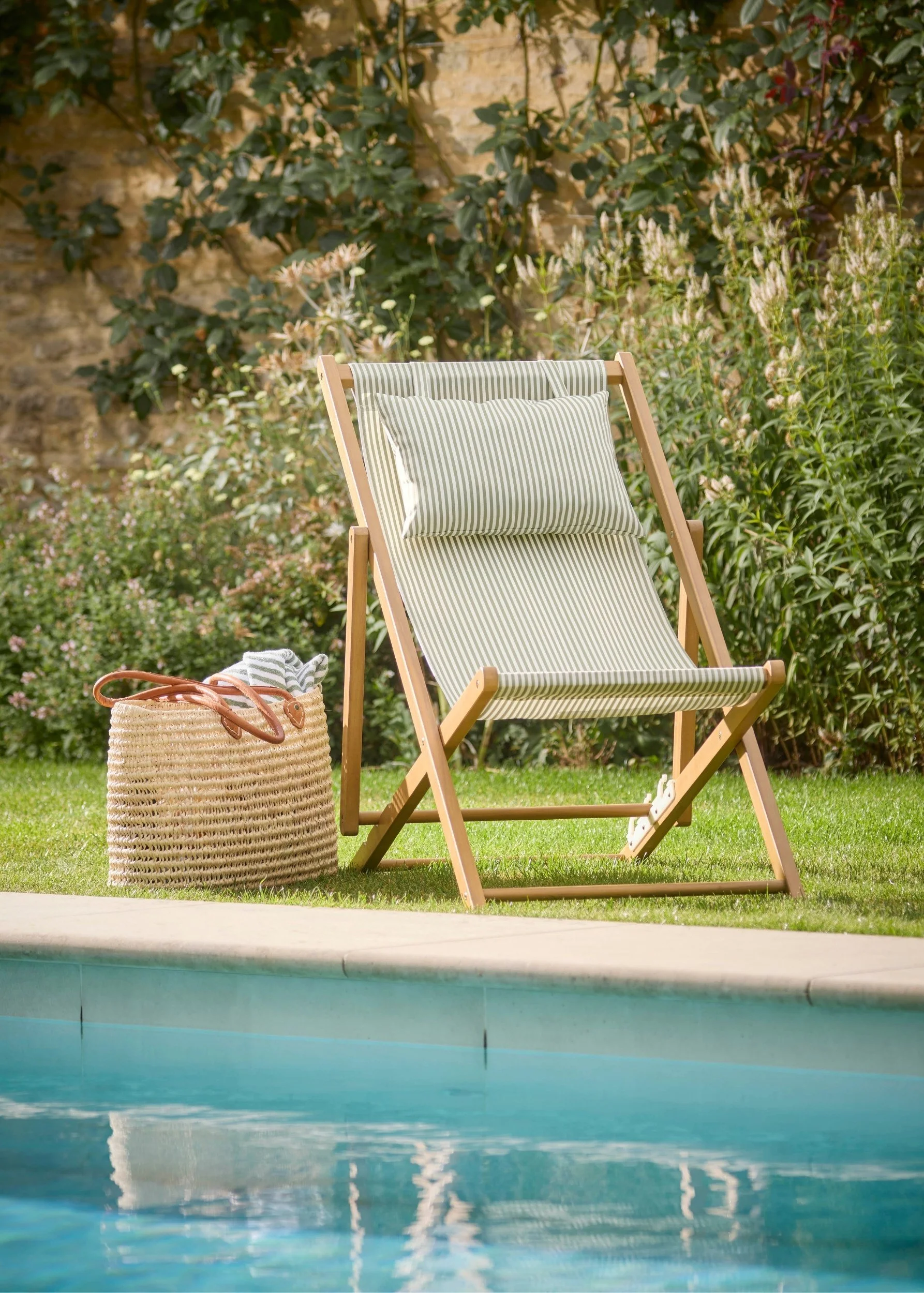 A stripy green deckchair with cushion and a rattan bag on the grass next to a swimming pool in front of a border of flowers