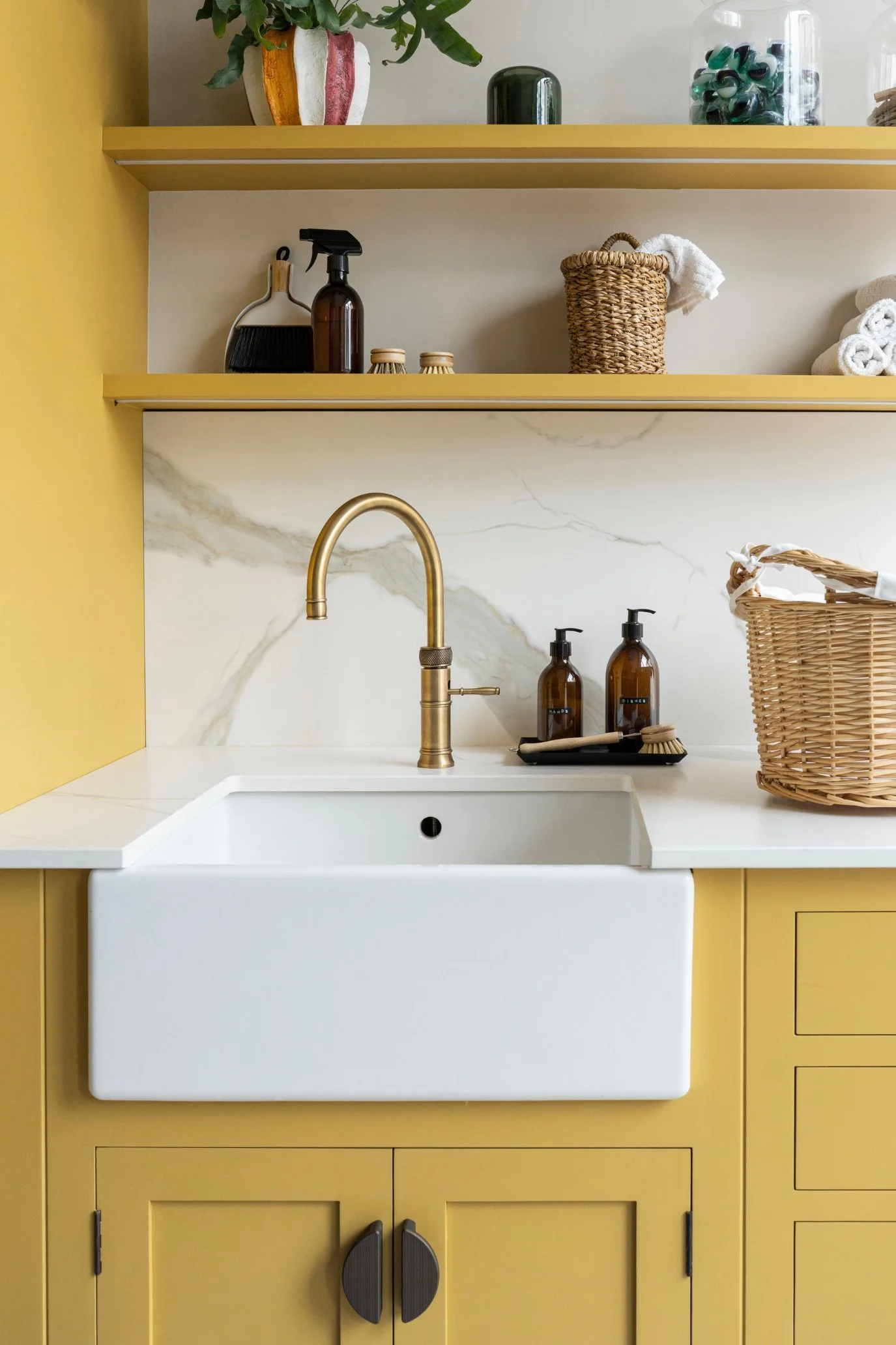 A kitchen with the cabinetry and shelves painted in a deep yellow and styled with plants, small wooden scrubbing brushes, baskets and bottles.