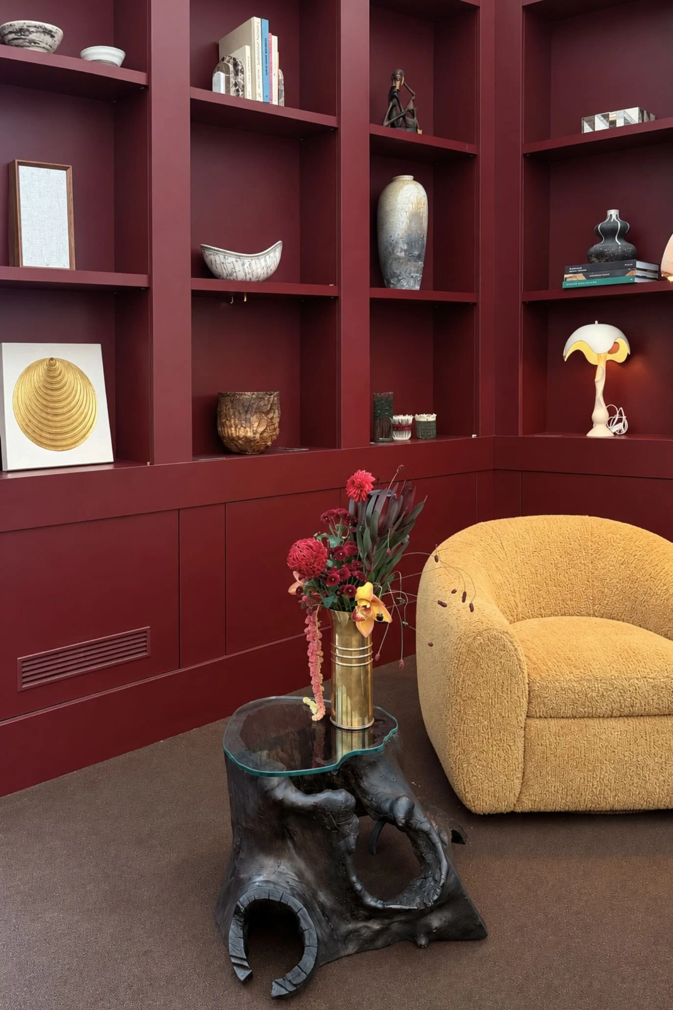 Wine-coloured shelving styled with ceramics, books and gold circular artwork beside a mustard armchair and sculptural table.