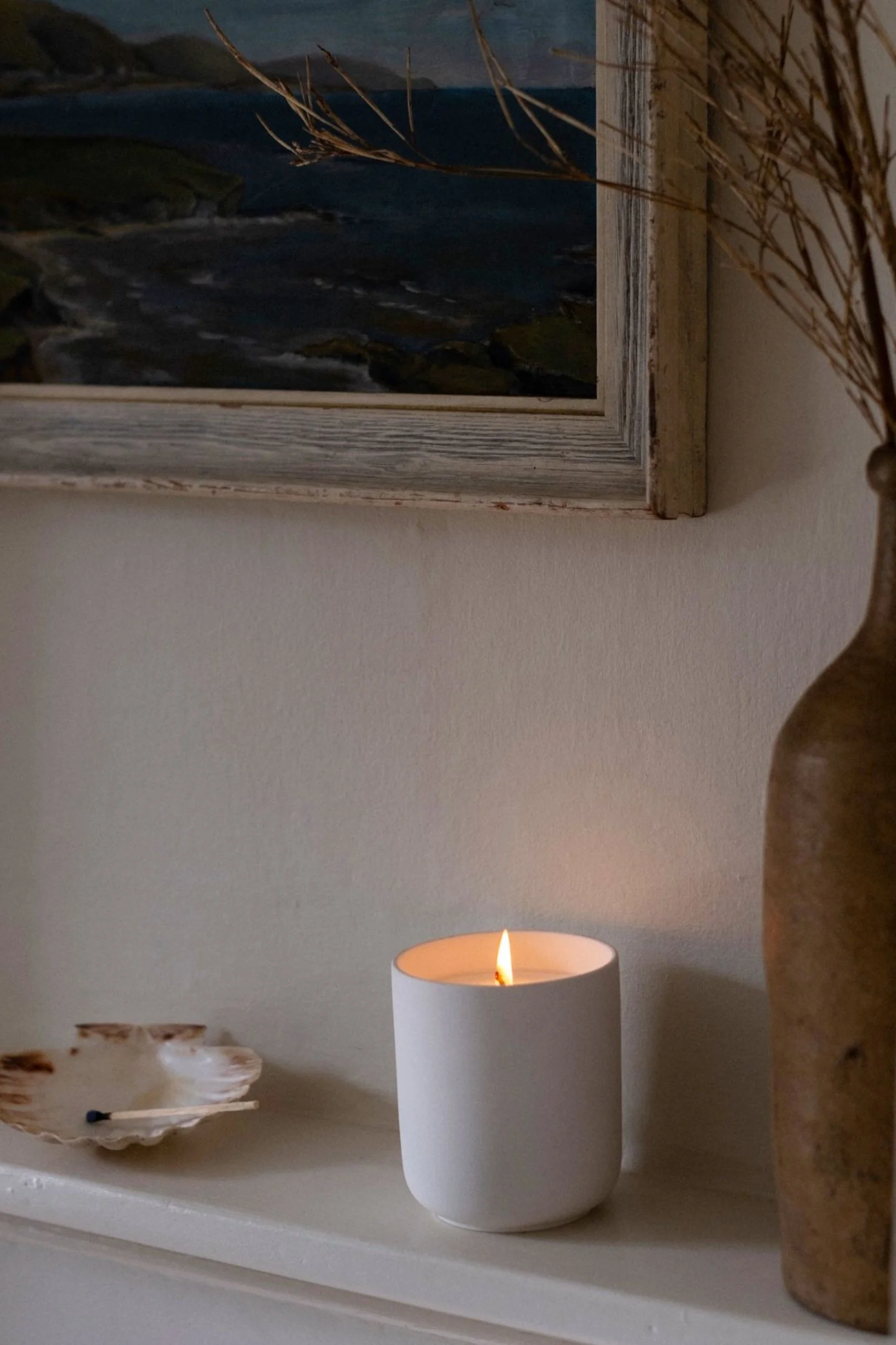 A white scented candle lit next to an old brown bottle, a shell with a match in it and a classic painting on the wall