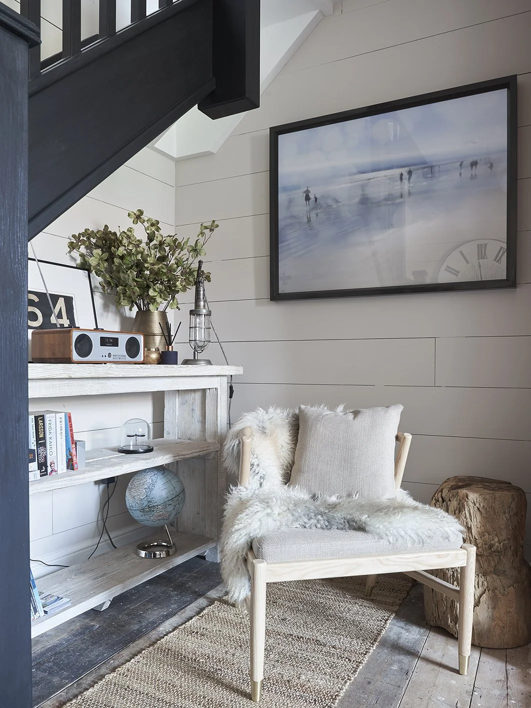 A chair with a sheepskin and cushion next to a shelving unit with a globe, radio, a light and a vase of dried leaves. A painting of a beach hangs above the chair