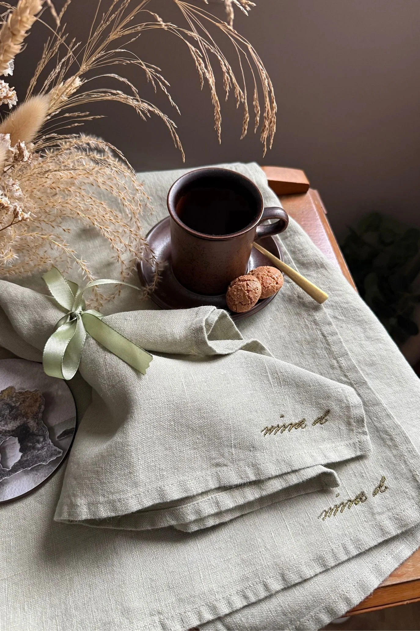 Sage green linen napkin tied with ribbon on a table styled with ceramic tableware and dried grasses.