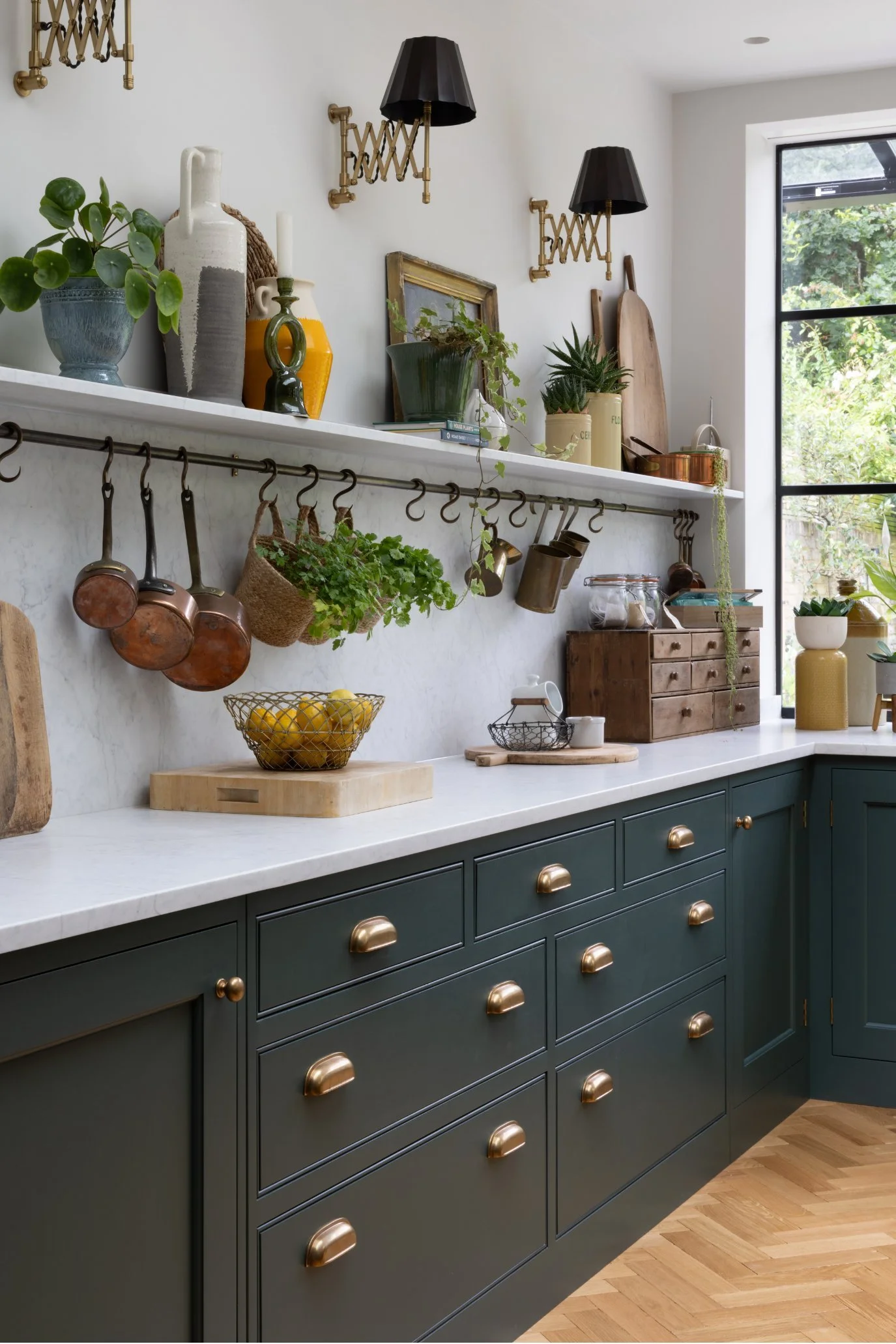 A kitchen in a rich deep grey with marble countertops, cplashback and a shelf which has been styled with plants, ceramics, chopping boards and a guilt frame.