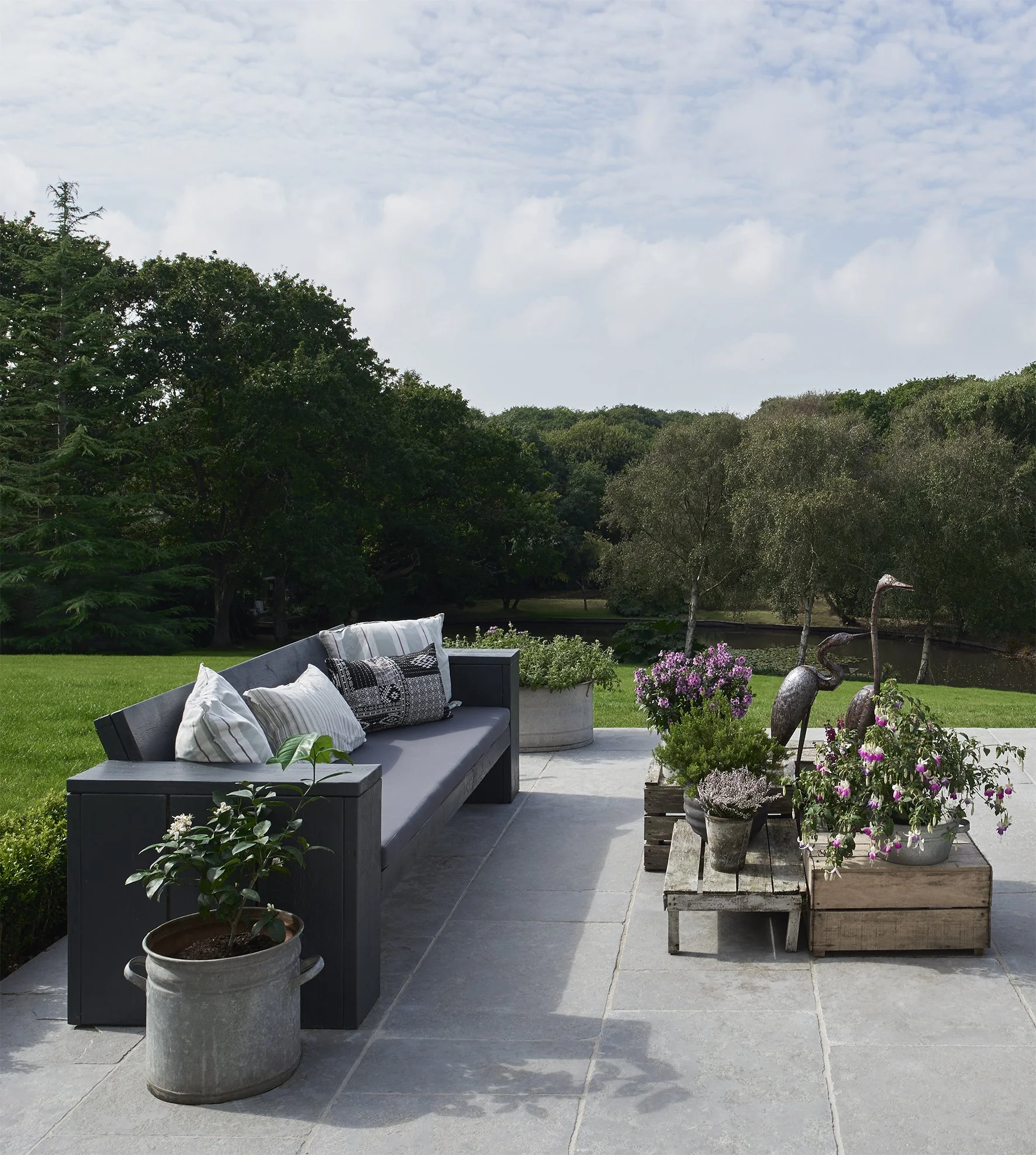 homemade furniture  made from scaffolding planks painted in dark grey with a mixture of grey and white linen cushions, flowers in planters with view of pond.