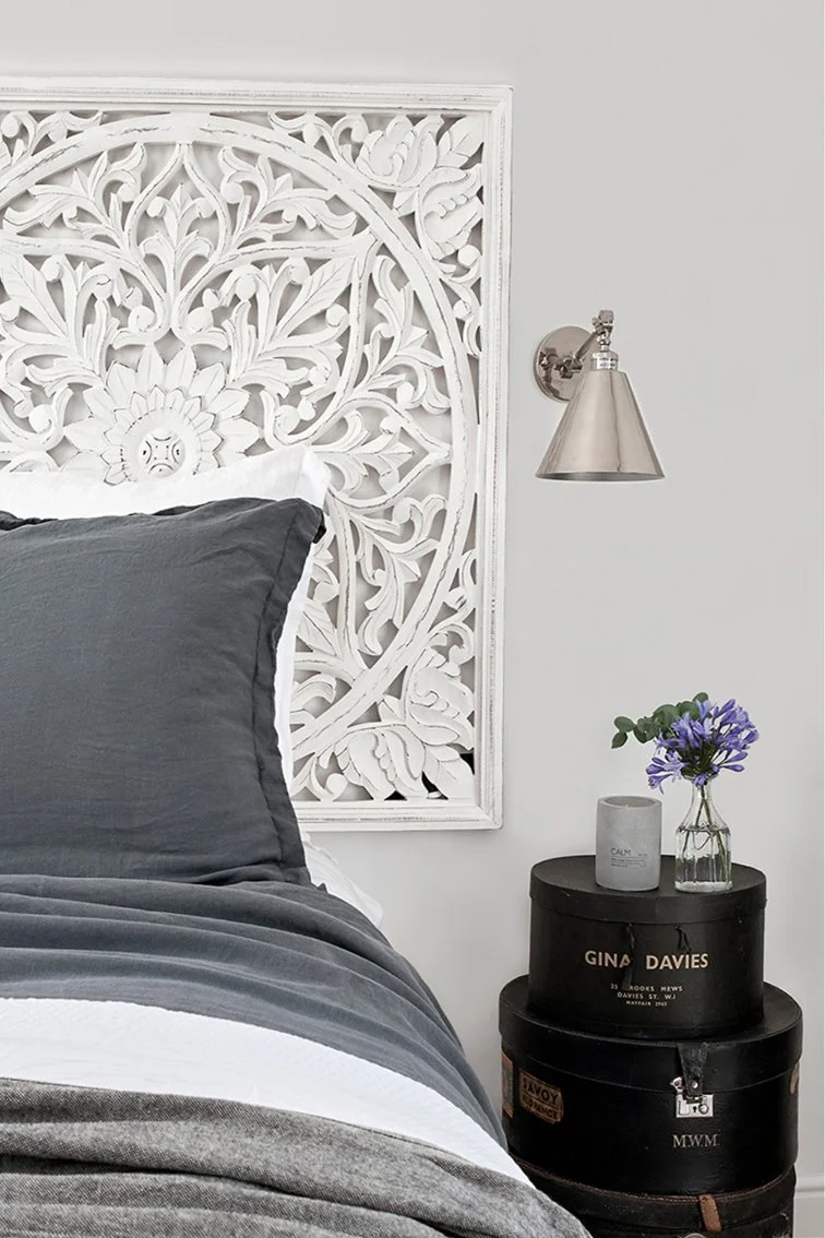 A bed with grey and white bedlinen with a decorative fame behind in white and a stack of three hat boxes as