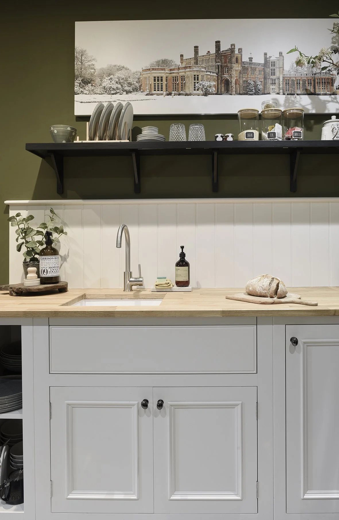 A white kitchen unit with a sink and pale wooden countertop with a sage green wall and painting of a castle in the snow above a shelf with plates and jars on it
