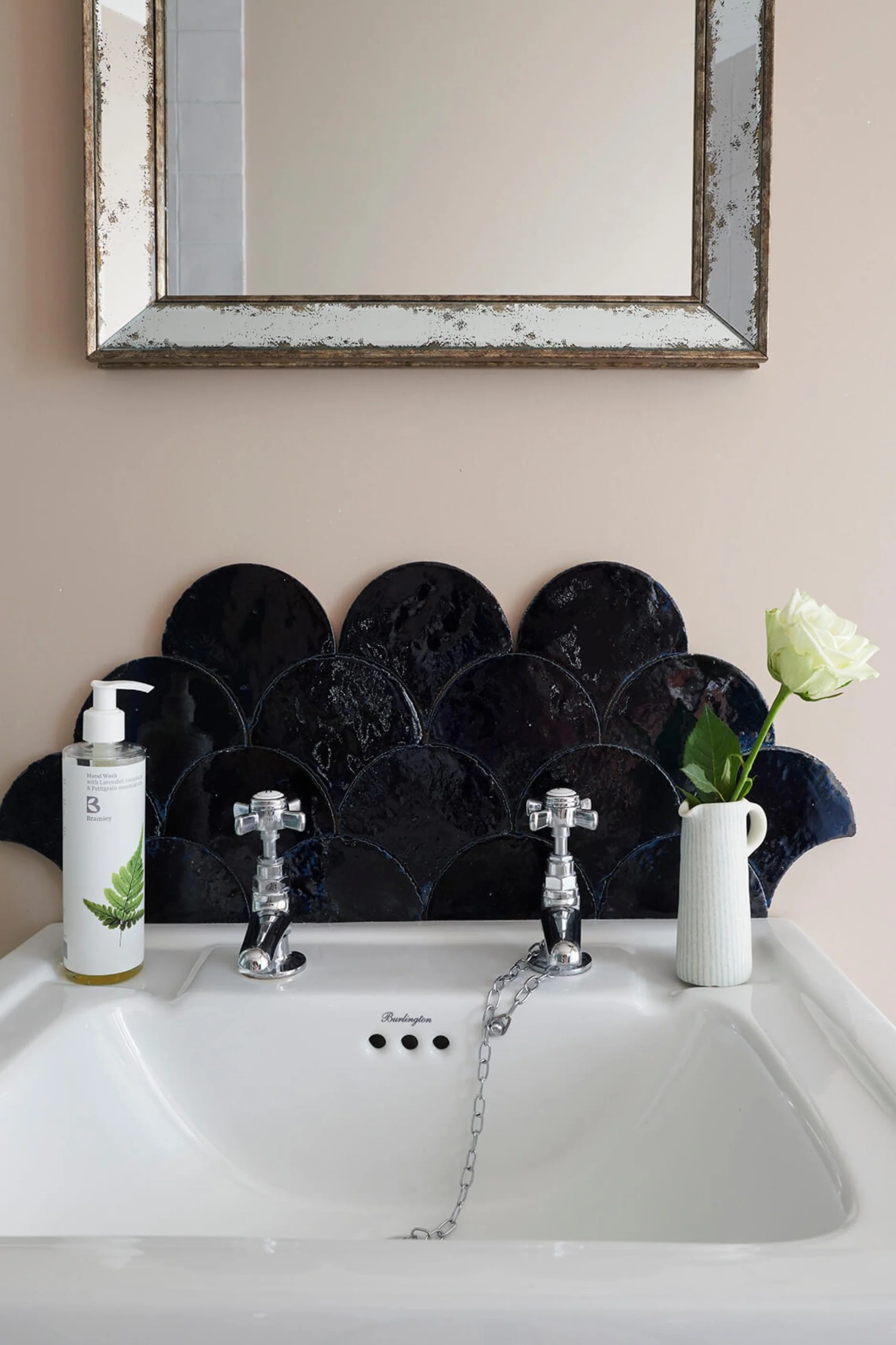 Close up of a basin with black glazed art deco style tiles, a proctor bottle, a rose in a mall white jug and a vintage style mirror hanging above