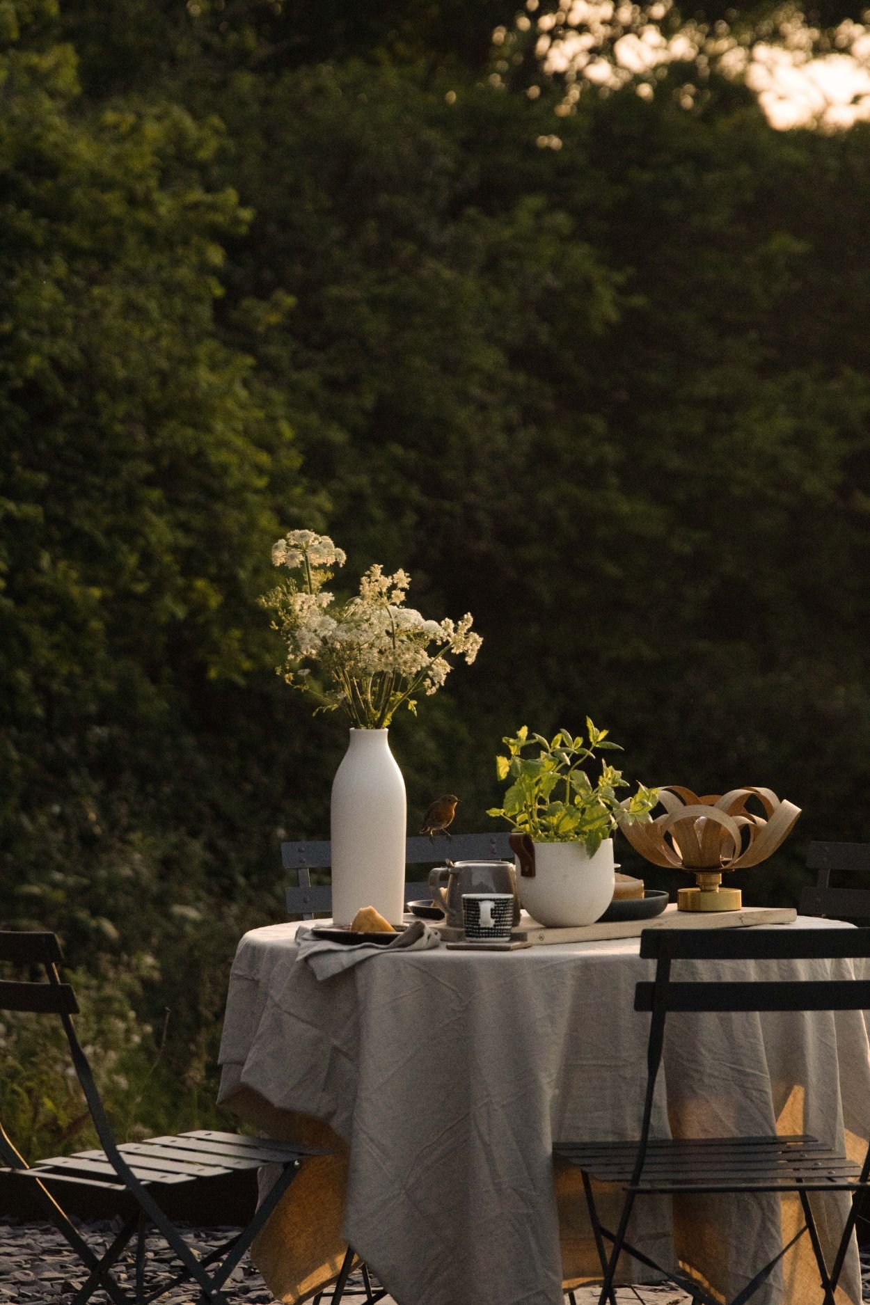 Outdoor dining table styled with a linen tablecloth, flowers, herbs and a portable lamp at sunset.
