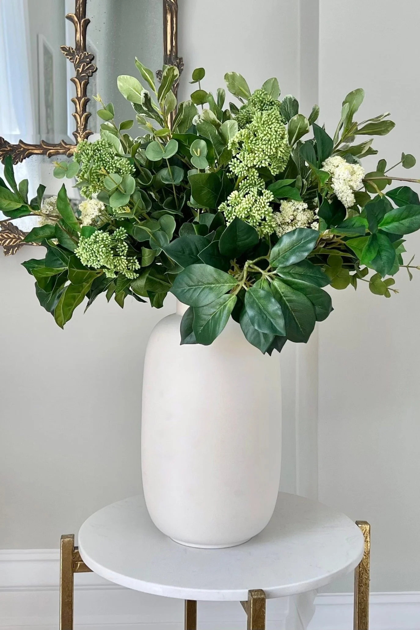Faux green foliage arranged in a tall white vase beside a decorative mirror.