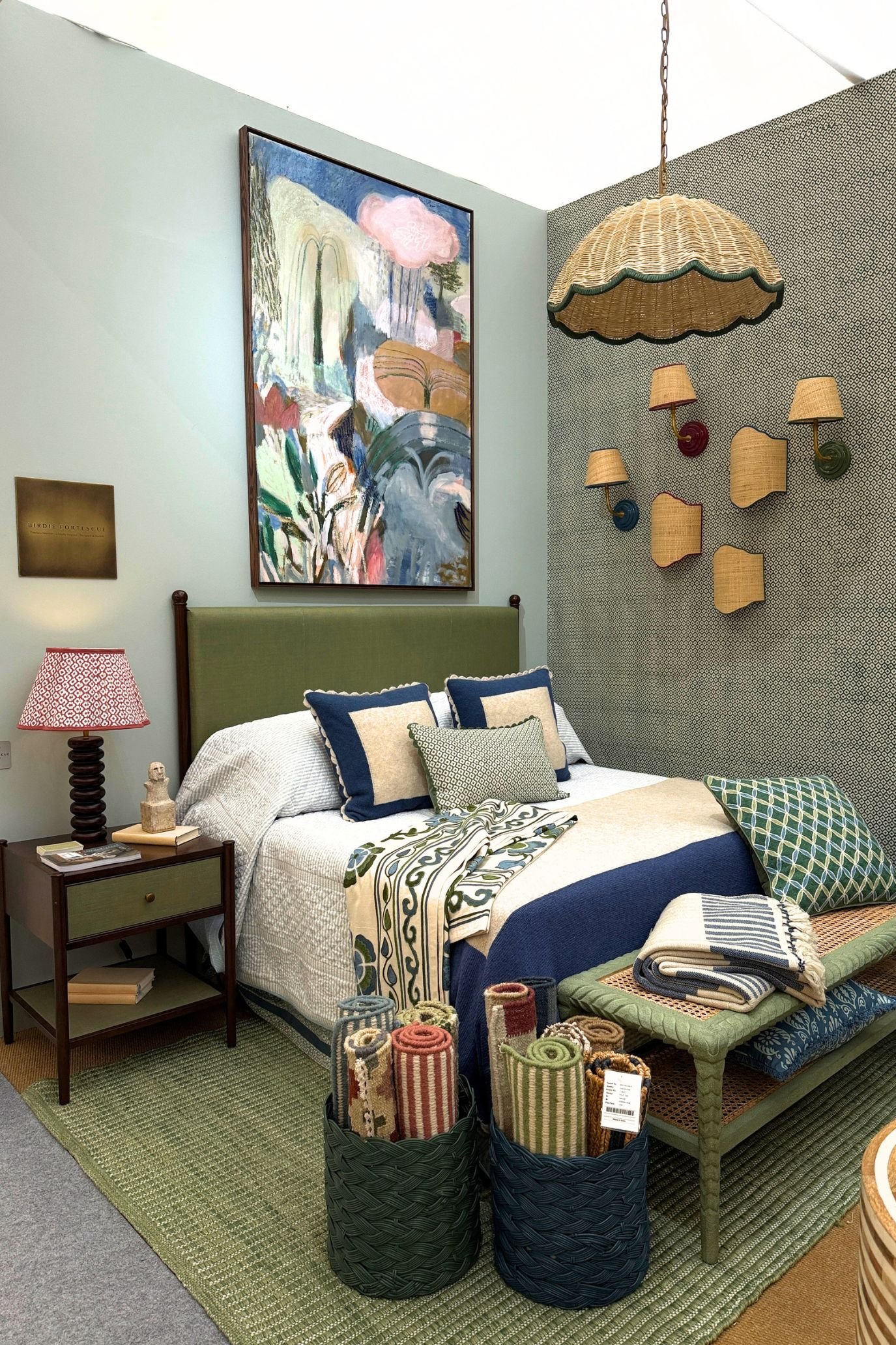 A bed with a green headboard and large artwork above next to a wall with a selection of rattan wall sconces and a pendant light