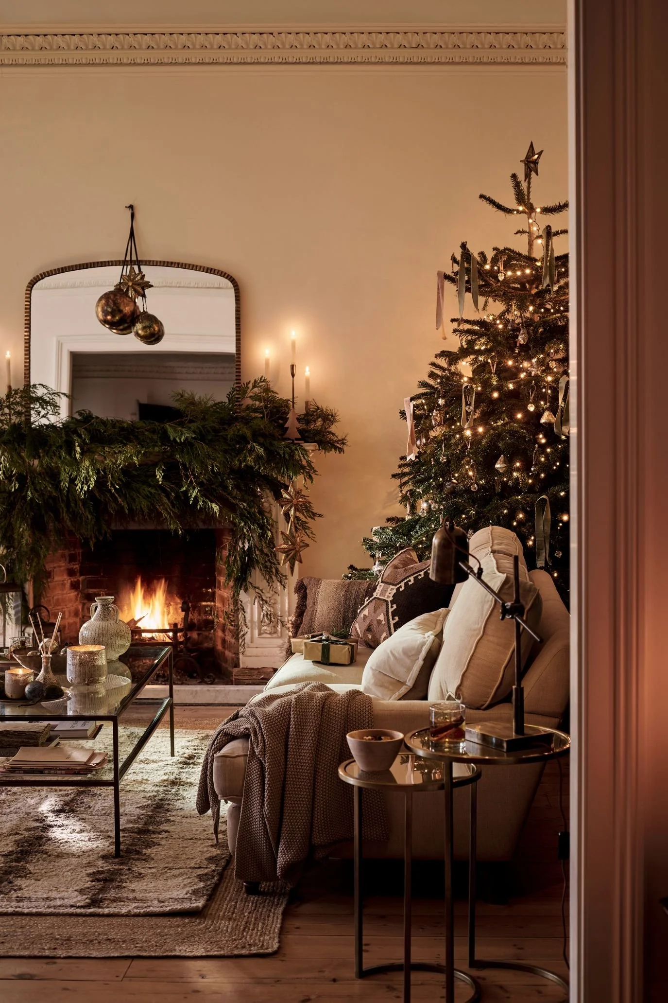 Christmas tree with softly glowing lights and muted ornaments beside a greenery-covered fireplace in a cosy country living room.