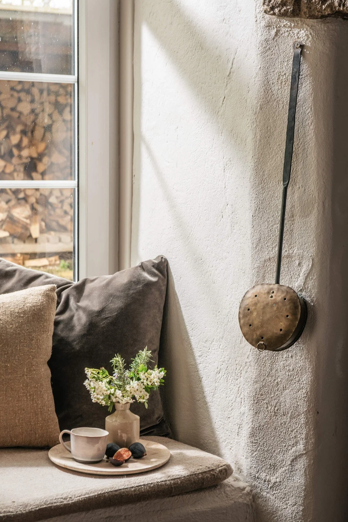 A rustic interior window seat with a brass bed warmer on the wall and cushions with a tray and earthenware on the seat