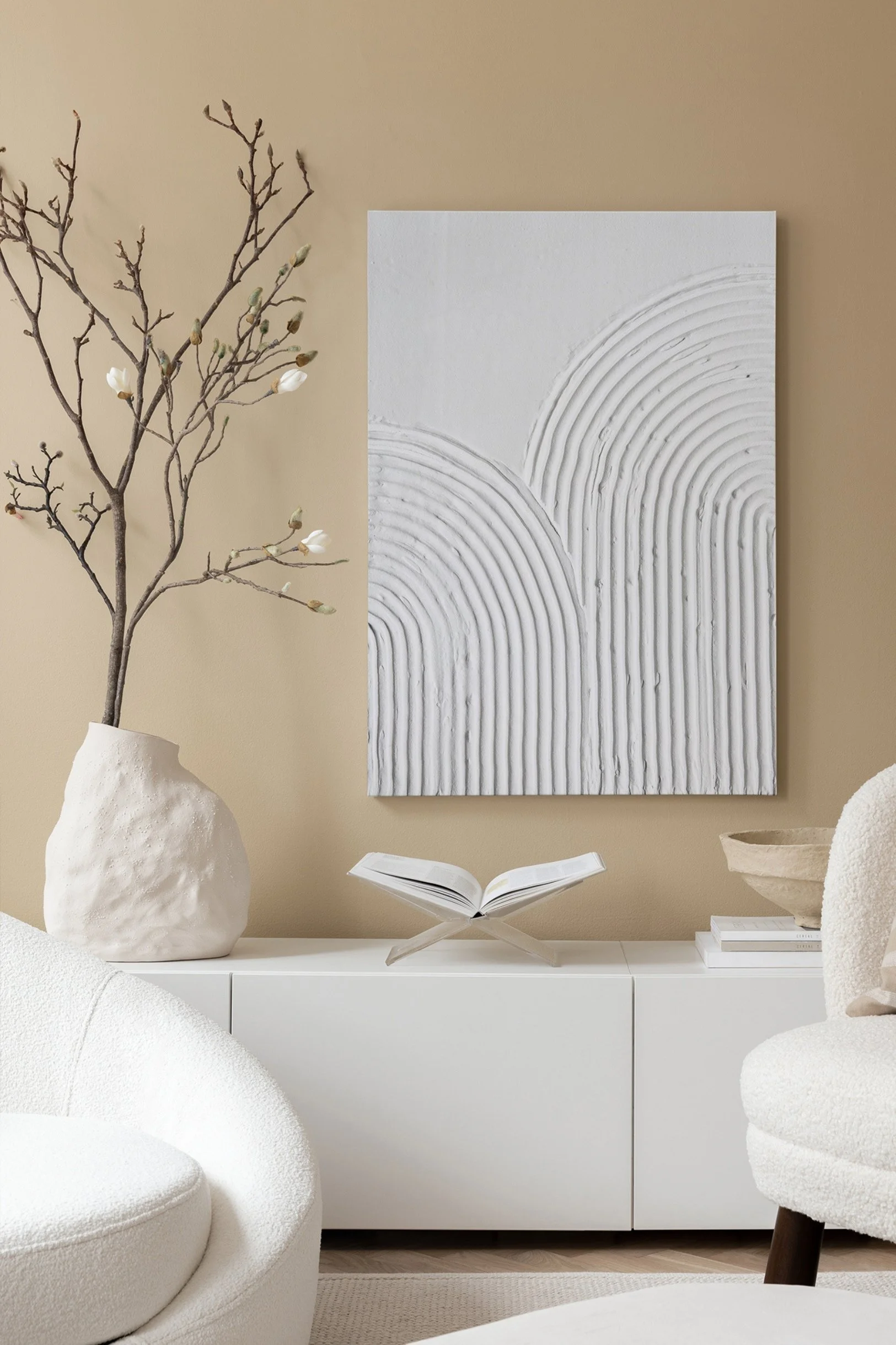 An abstract white canvas on a beige wall above a white cabinet with a textured vase and branches