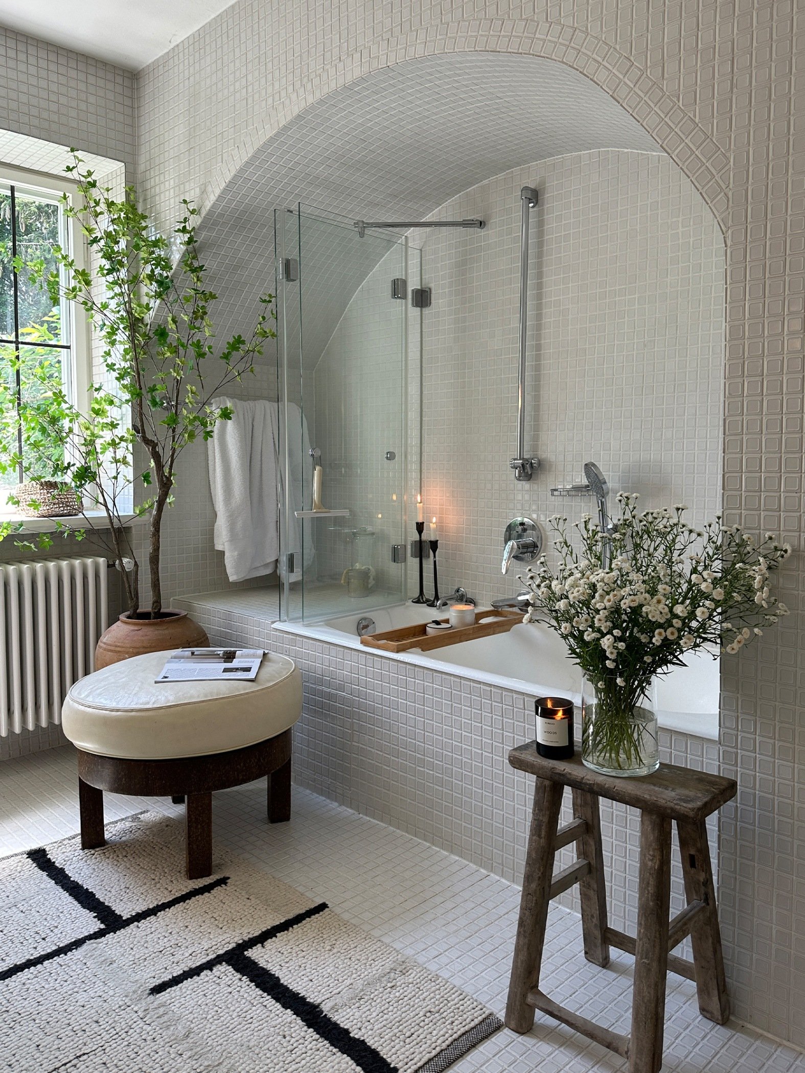 A bathroom with an arch shaped wall and bath with grey tiling. There is a wooden stool with flowers and small ottoman with fabric in cream on top
