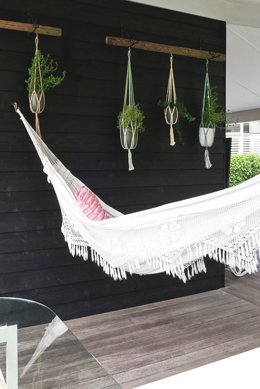a macrame hammock hanging from a black wooden exterior wall next to four plants in hanging macrame pots.