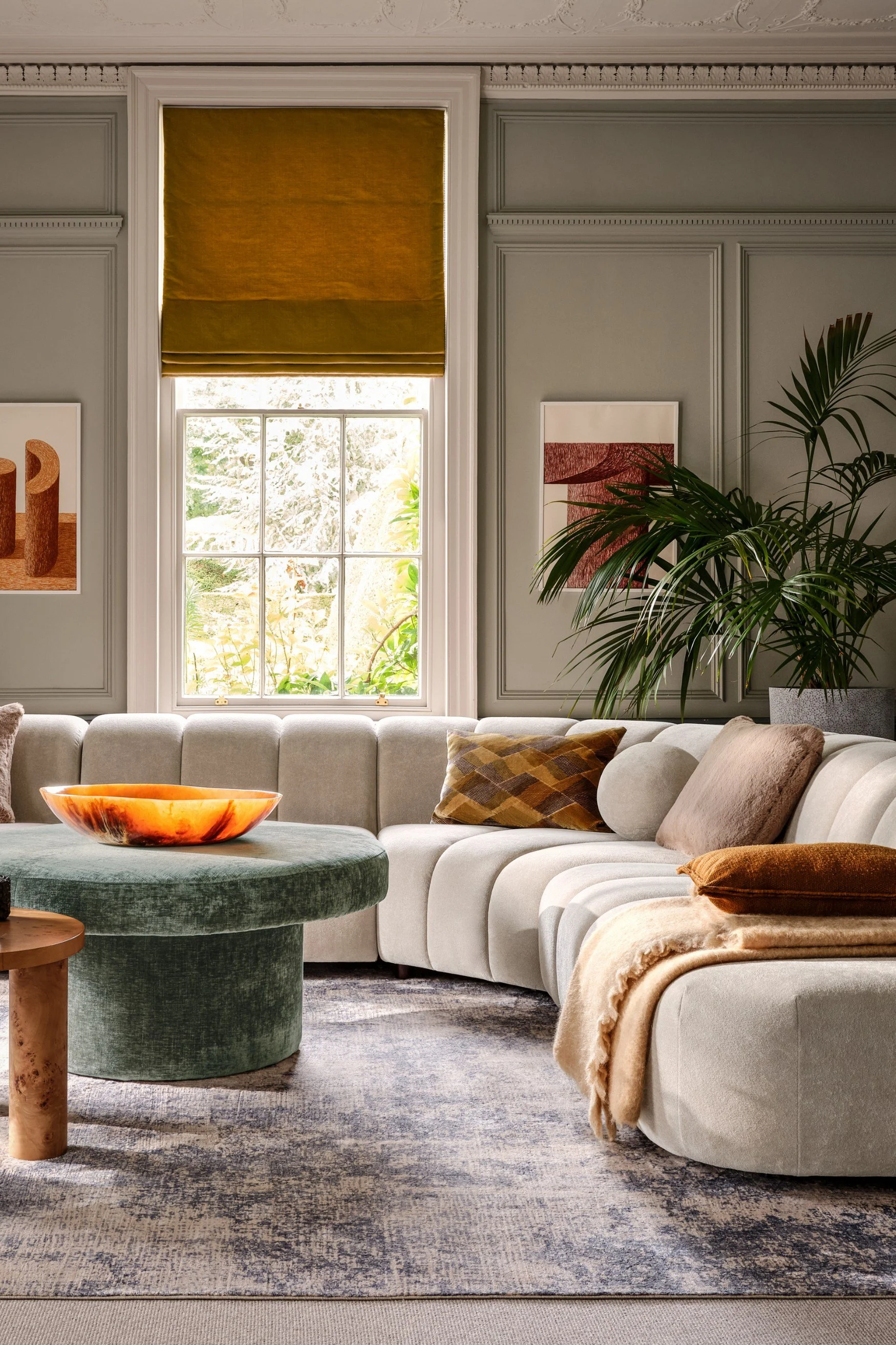 A living room with a large cream circular sofa with ochre toned cushions and a plant behind. There is a green velvet table with a stone yellow dish on it