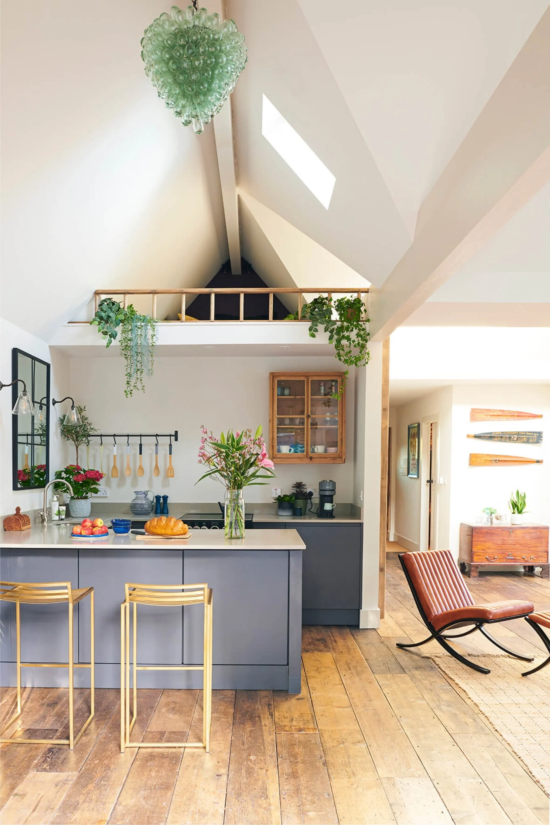 An open plan kitchen and living area with a vaulted being and a glass pendant hanging from it. There are two brass bar stall, a windowpane mirror and a wood glass-fronted cabinet on the wall