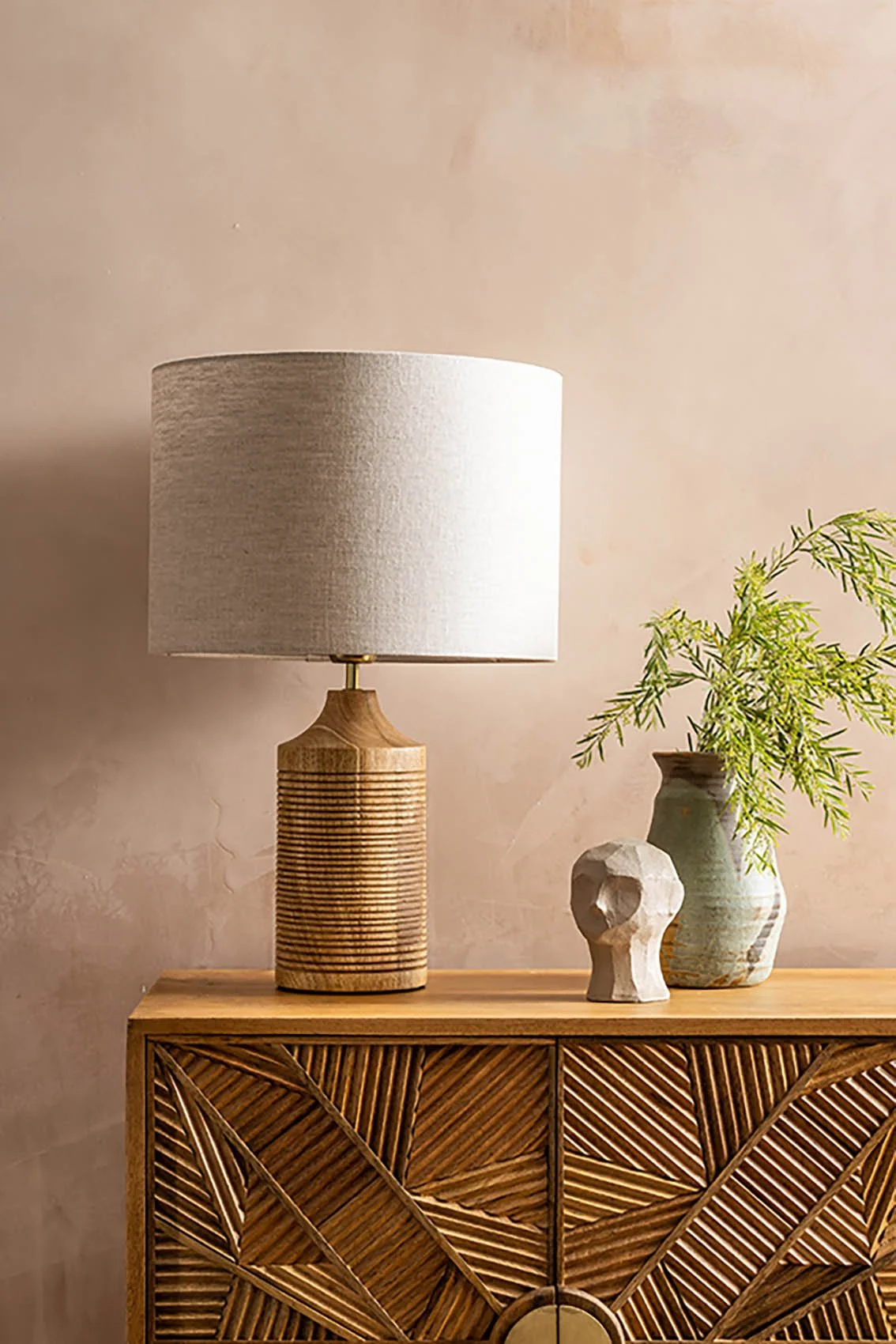 Home Decor items a wooden ribbed lamp with beige linen shade on a  low cabinet with a face sculpture and vase with greenery.