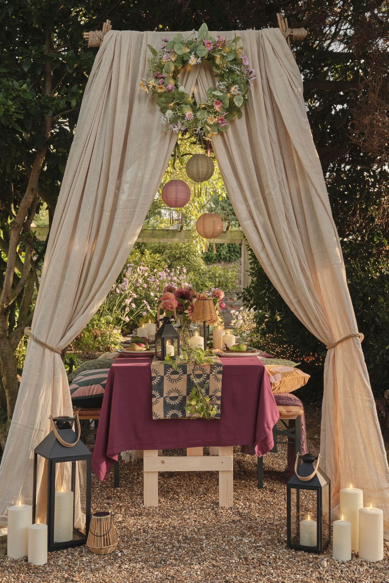 Outdoor dining table dressed with linens, lanterns and candles beneath flowing fabric drapes in a garden setting.