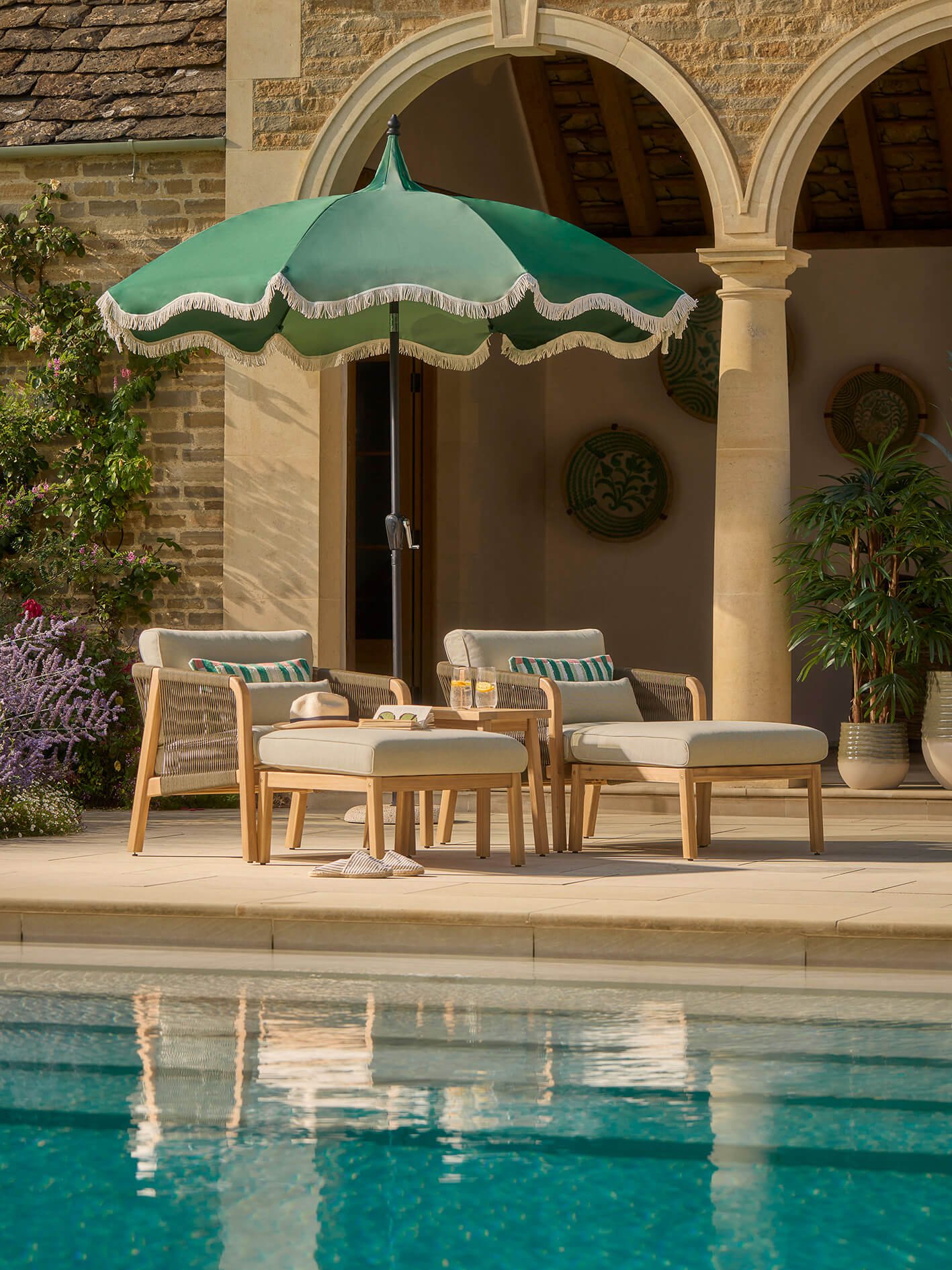 two wooden outdoor days beds with beige base cushions and green stripy cushions to match a green parasol by a pool