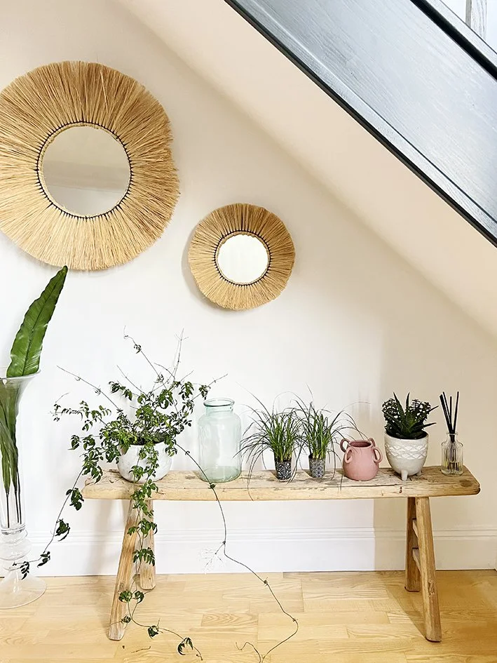 mirrors with straw in two different sizes under staircase and above a rustic wooden bench with plants and vessels.