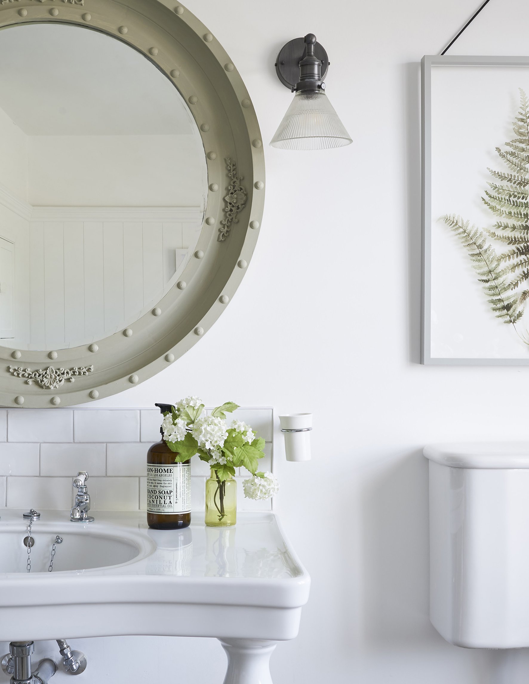 A bathroom basin with a product bottle nd small glass vase with flowers. There is an ornate round mirror above