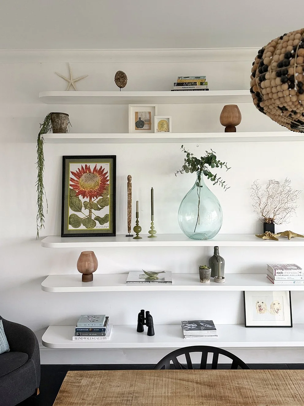 A set of floating shelves with art, candles, lamps, vases and plants with a wooden table in front