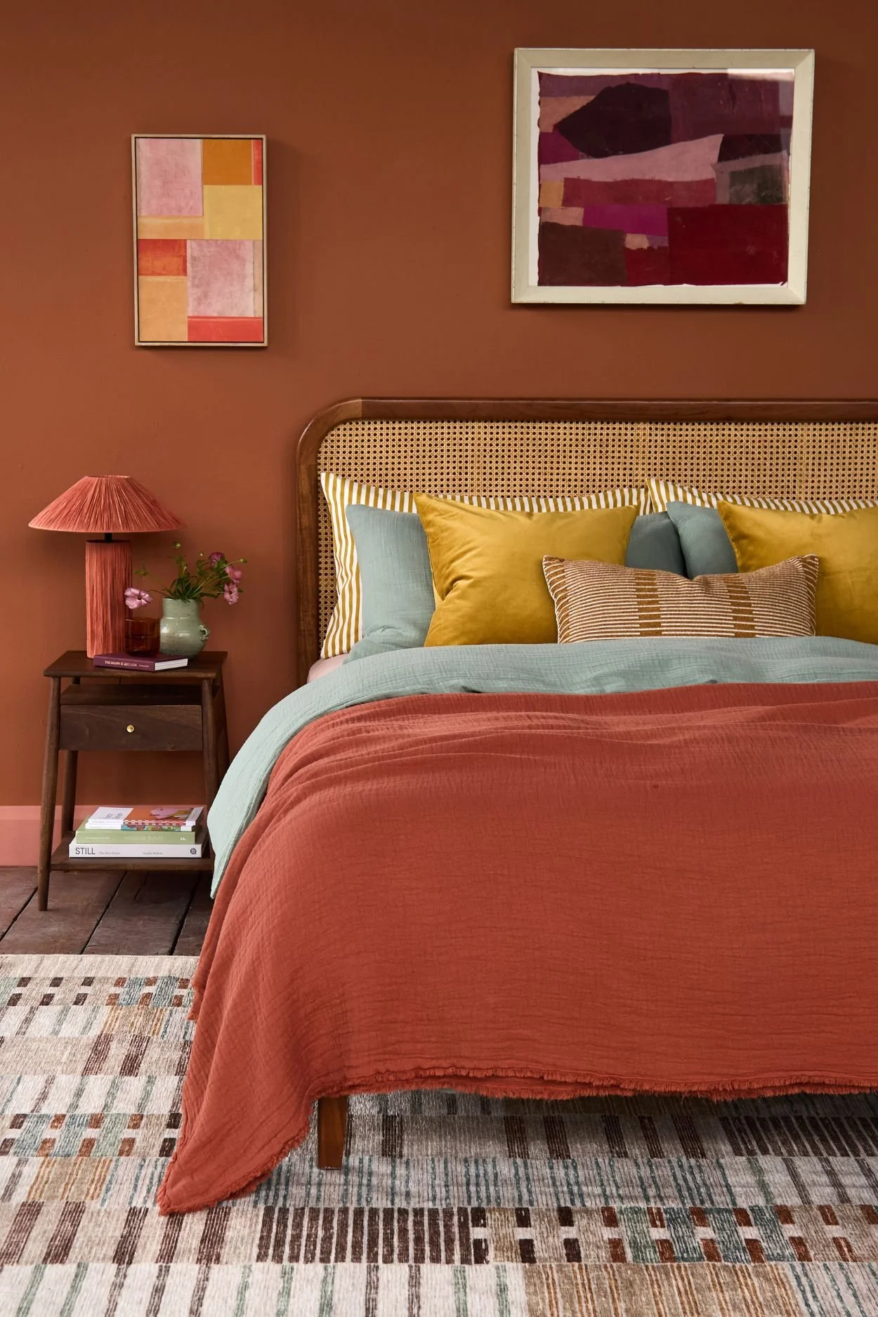 A rattan and dark wood bed dressed with blue and gold cushions and pillows and a terracotta throw that pairs with the wall in the same tone