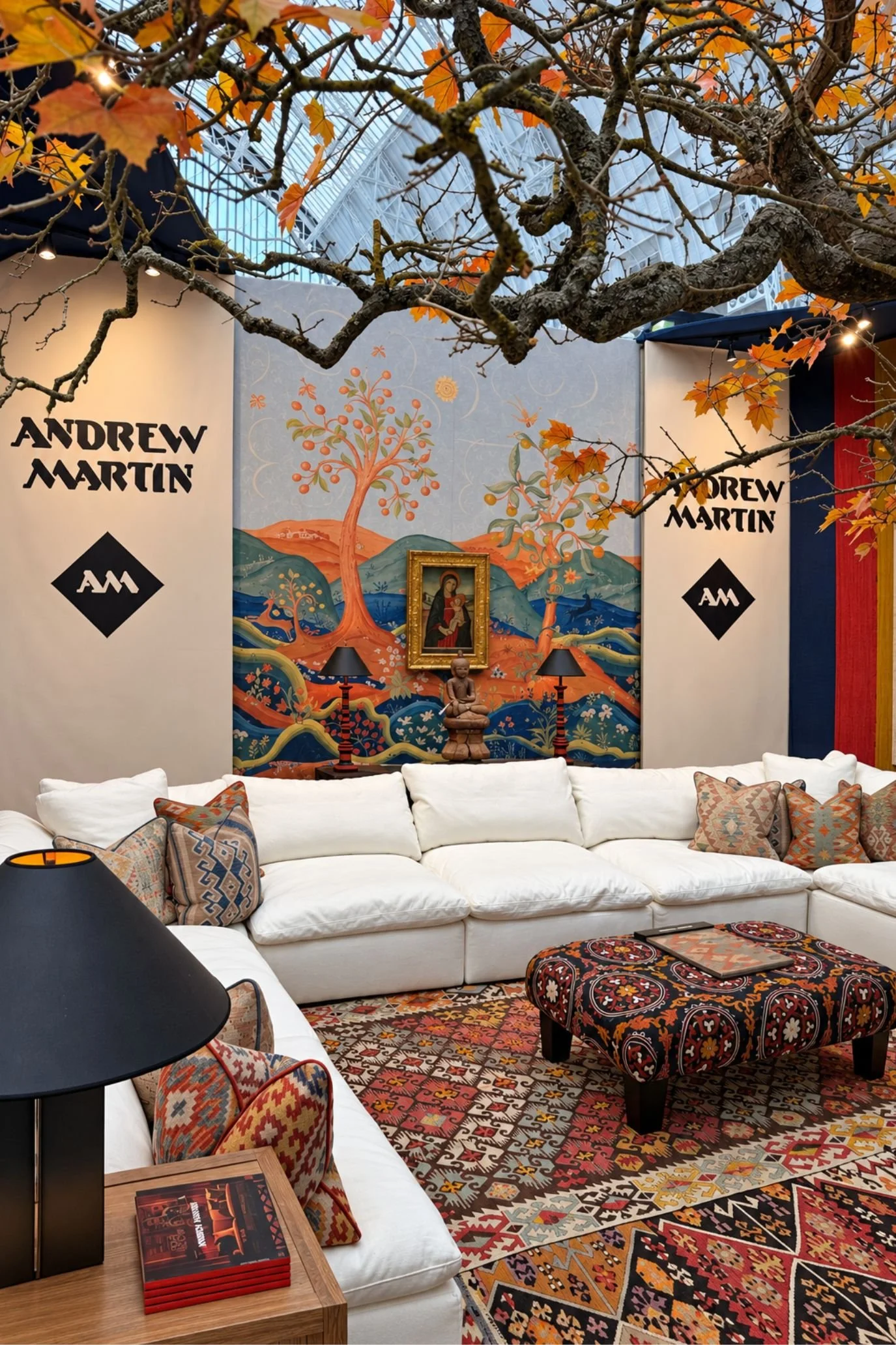 Andrew Martin stand with a white sofa, layered patterned rugs and kilim-style upholstery beneath a decorative tree canopy.