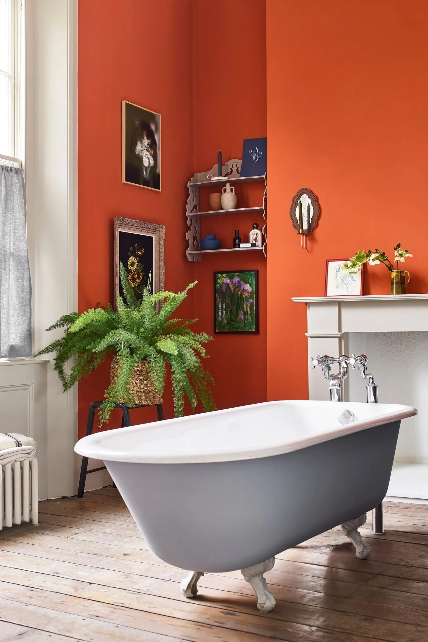 a roll top bath in grey infant of a fireplace and vivid orange wall which is decorated with a few paintings and a small vintage shelving unit.