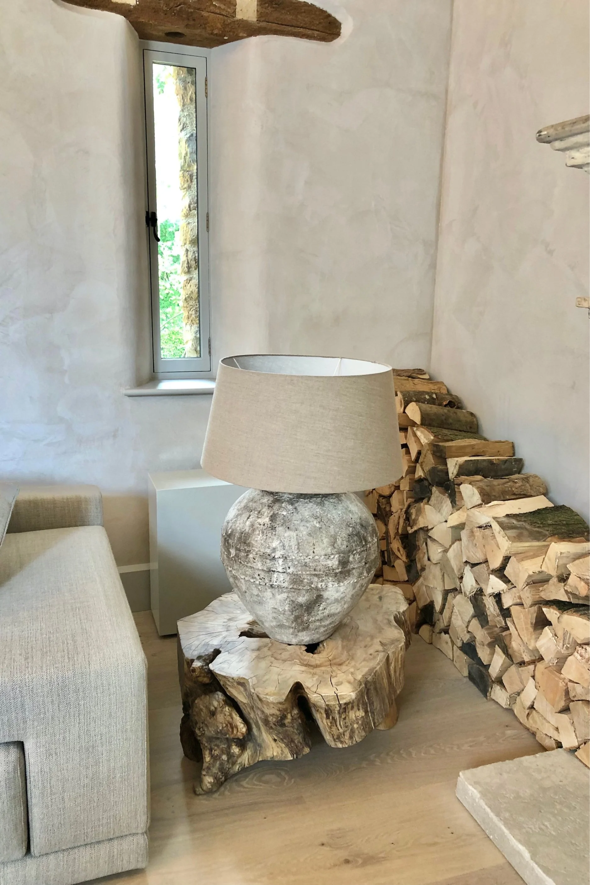 A lamp with a distressed base and cream linen shade on a wooden table made from a tree trunk. There is a pile of firewood piled up behind and a small narrow window