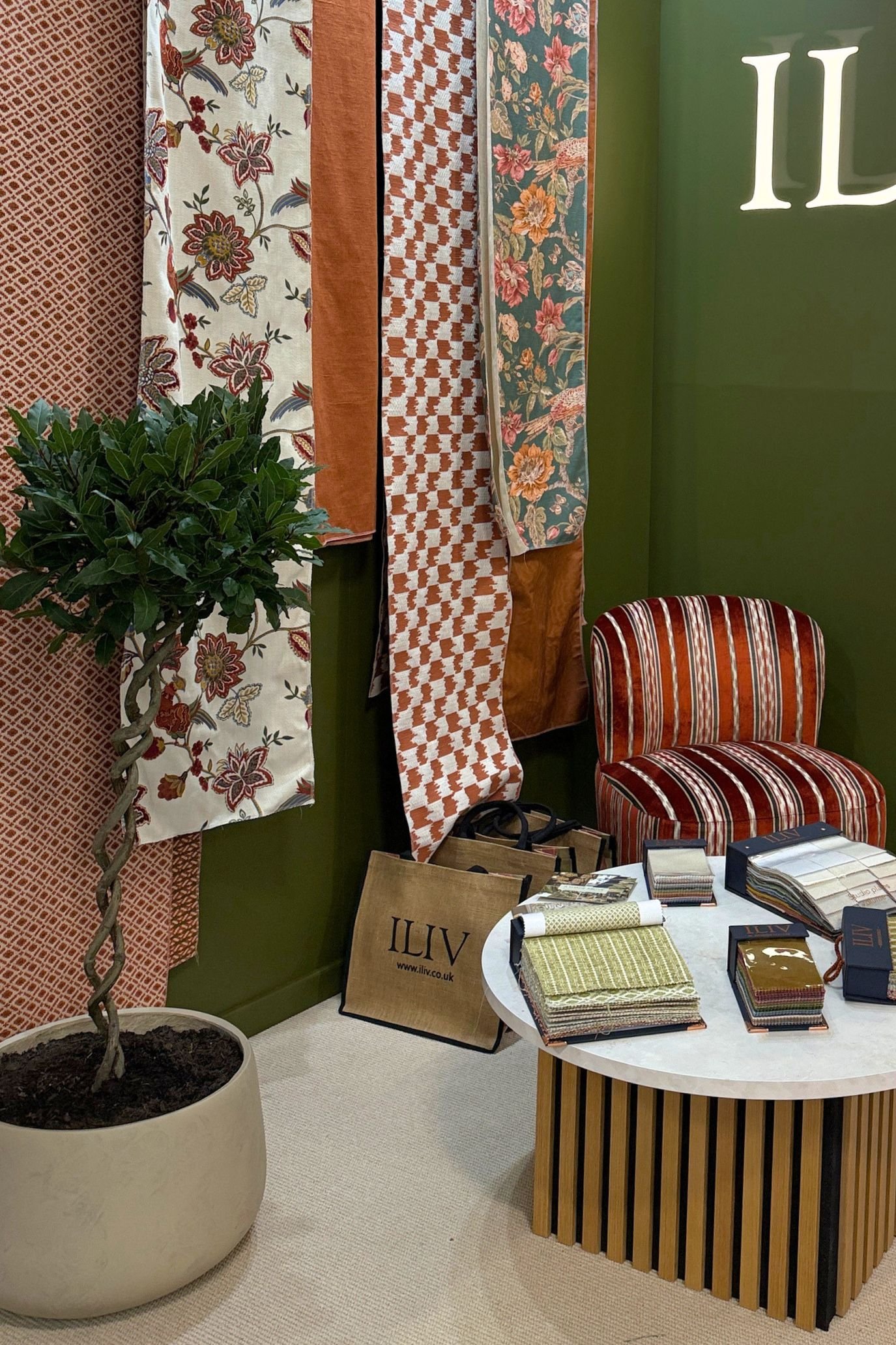 Floral, checked and striped fabrics displayed against green walls on the ILIV stand, beside a striped upholstered chair.