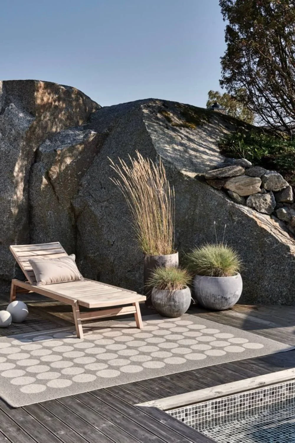 a grey rug with white stripes on wood decking by a pool with a wooden sun lounger and concrete pots with plants