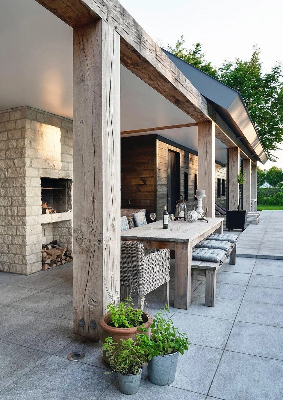 An outdoor long terrace with a beamed wood covered area and a dining table and chairs next to a built in open oven.