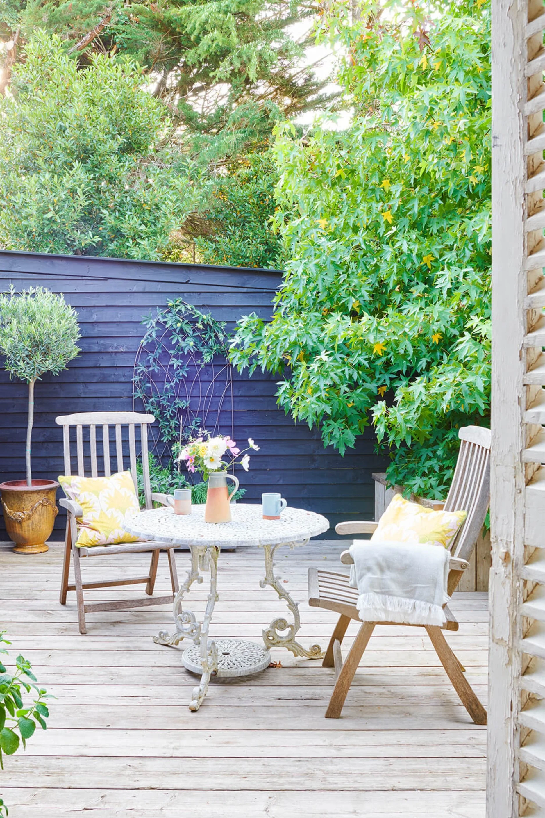 A decked courtyard with a white wrought iron table with a jug of flowers and two cups. There are two wooden chairs with yellow cushions and a throw