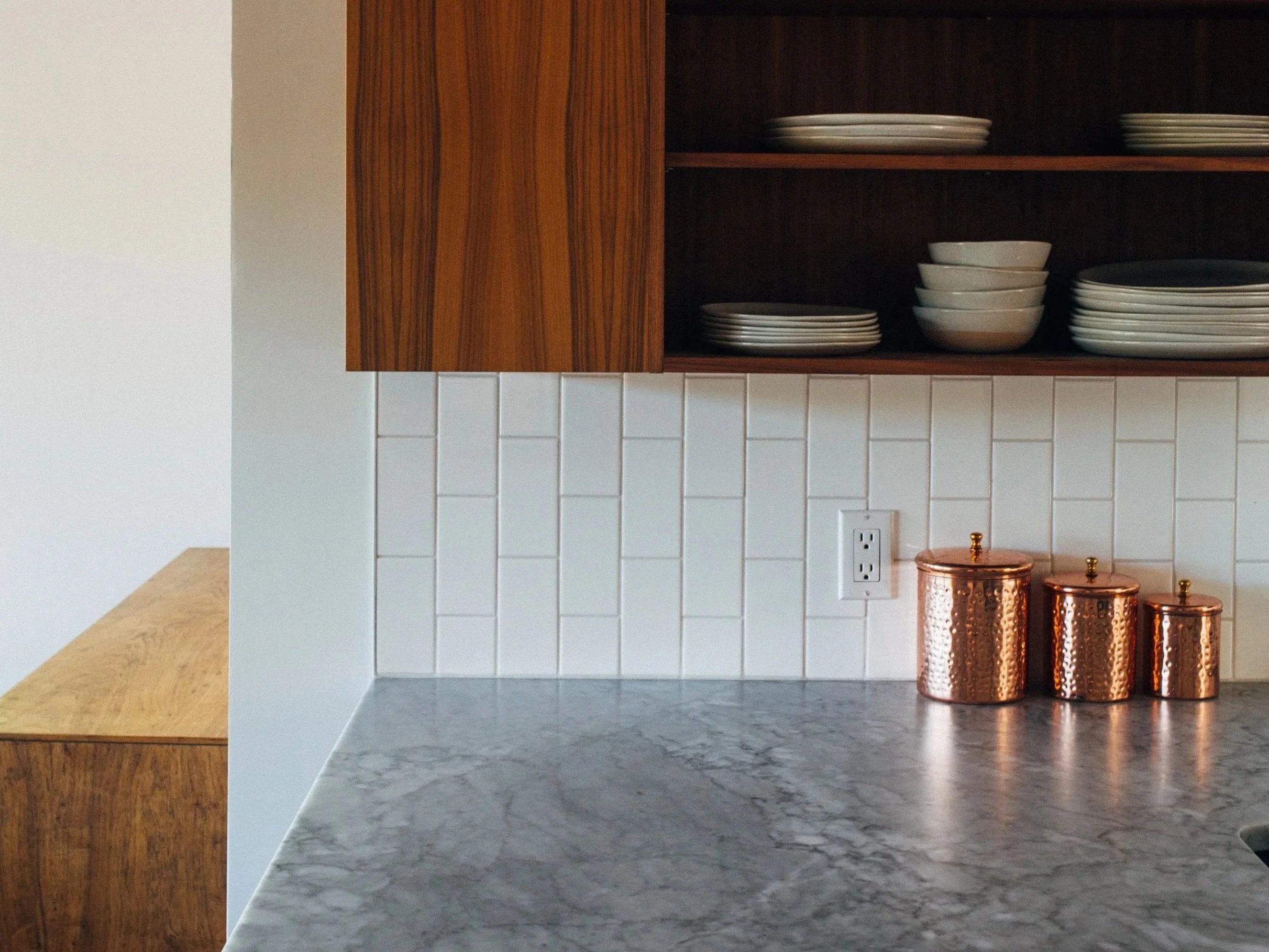 Modern kitchen detail featuring walnut cabinetry, open shelving with dishware, white tile backsplash, and a stone countertop.