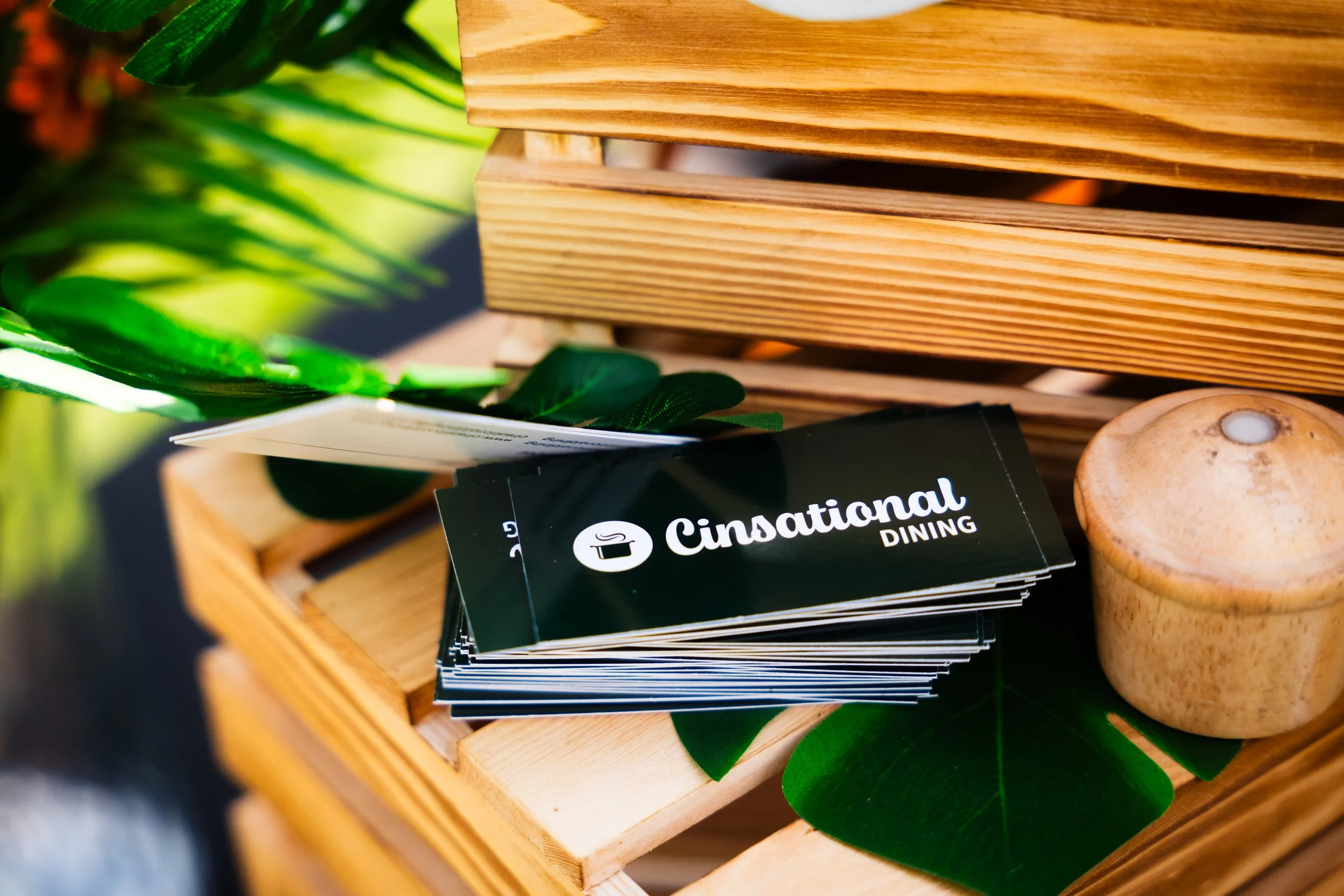 Close-up of a wooden tray holding black cards with white text reading 'CinationaL DINING' and a cooking pot icon, surrounded by green leaves and a small beige mushroom.