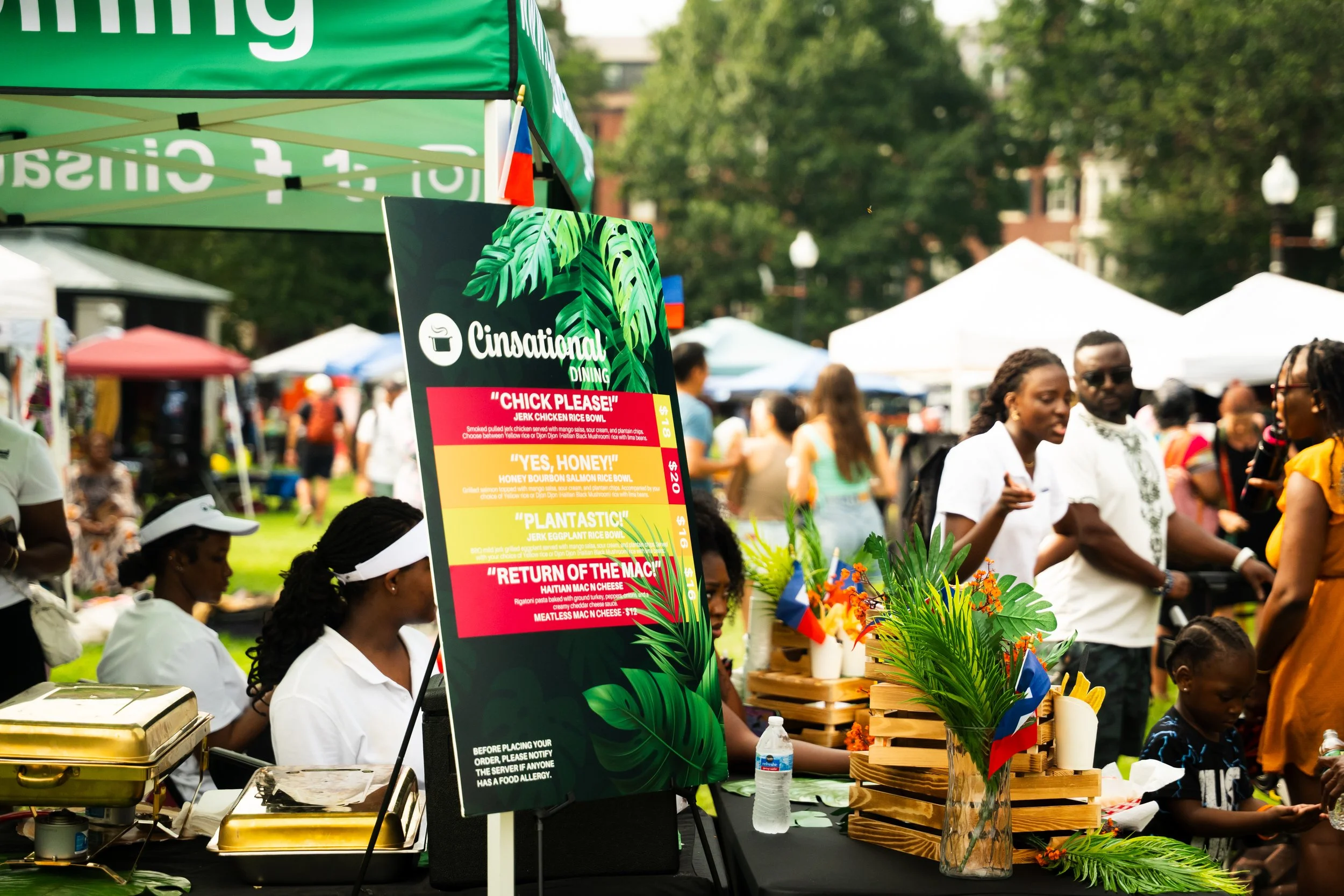 A lively outdoor market with vendors and shoppers, featuring a green sign for CinationaL Dining, displays of food items, flowers, and small flags, with numerous people walking and browsing in the background.
