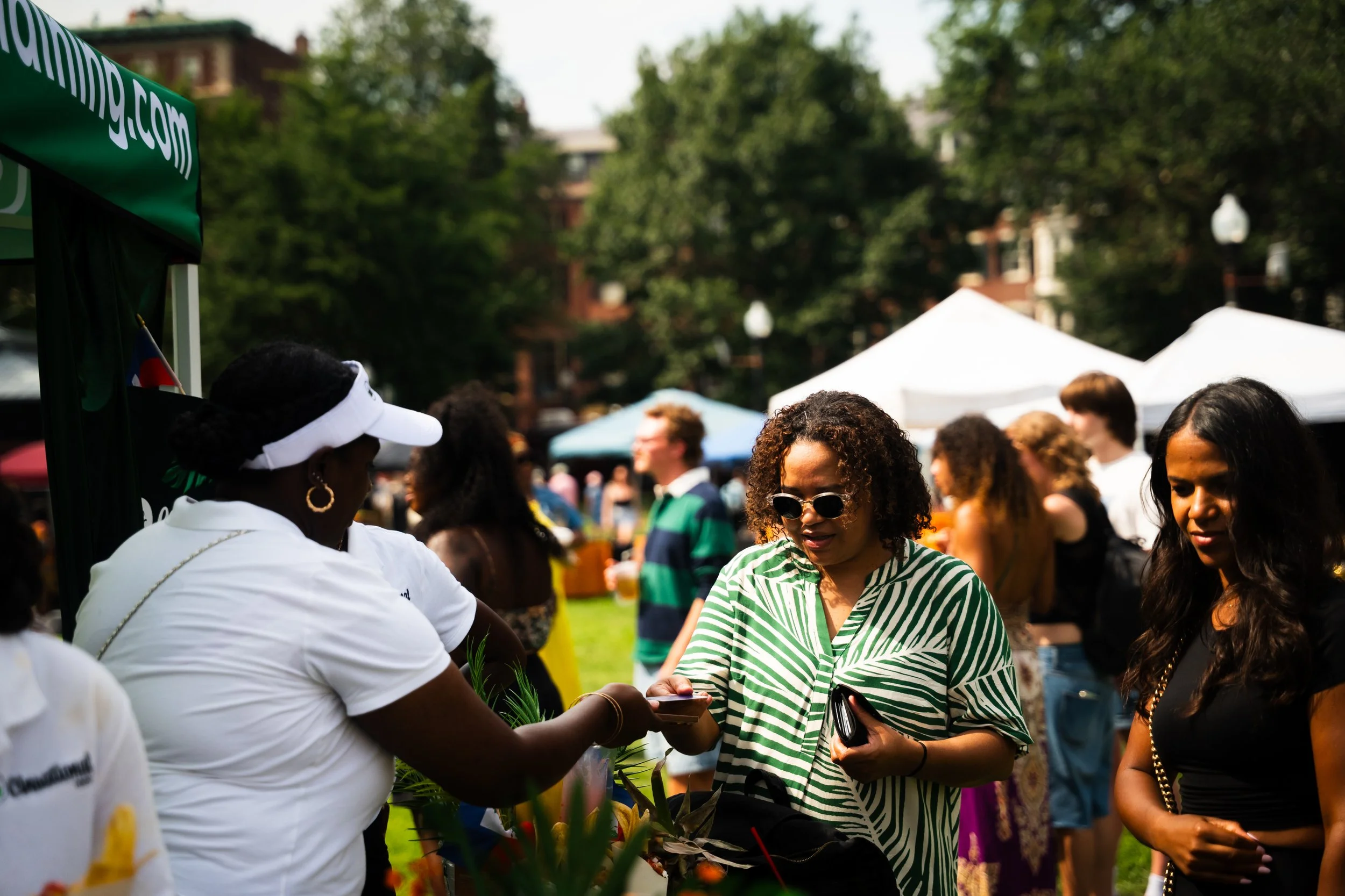 People gathered outdoors at a festival or event, talking and interacting under tents with trees in the background.