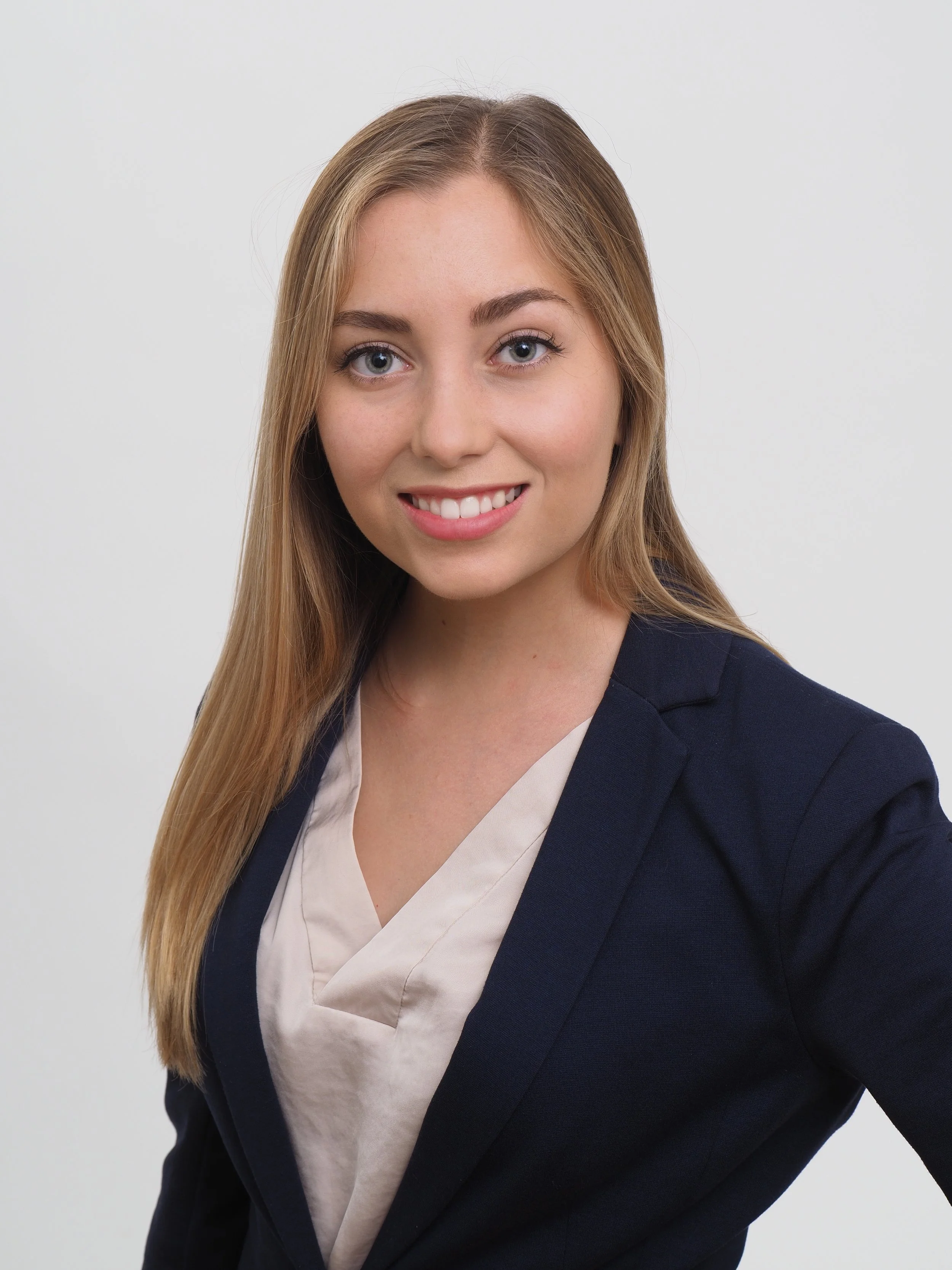 Professional woman with long blonde hair, smiling, wearing a navy blazer over a beige top, standing in front of a plain white background.