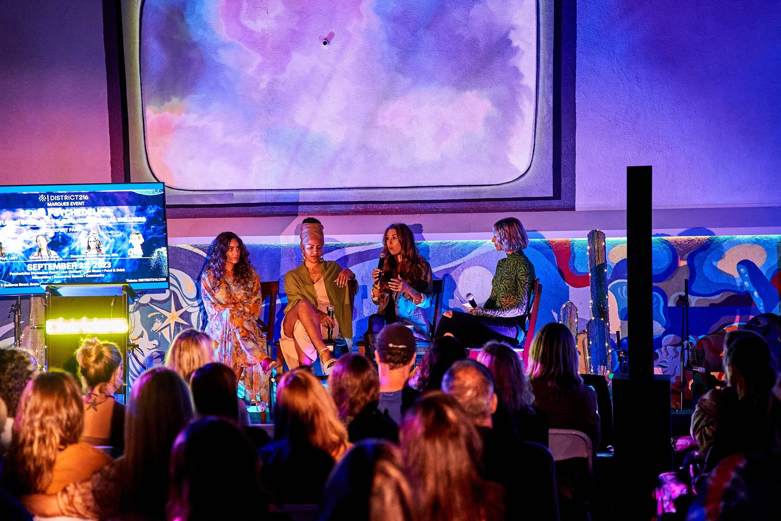 A panel discussion with four women on stage in front of an audience, with a colorful mural backdrop and a large screen showing event information.