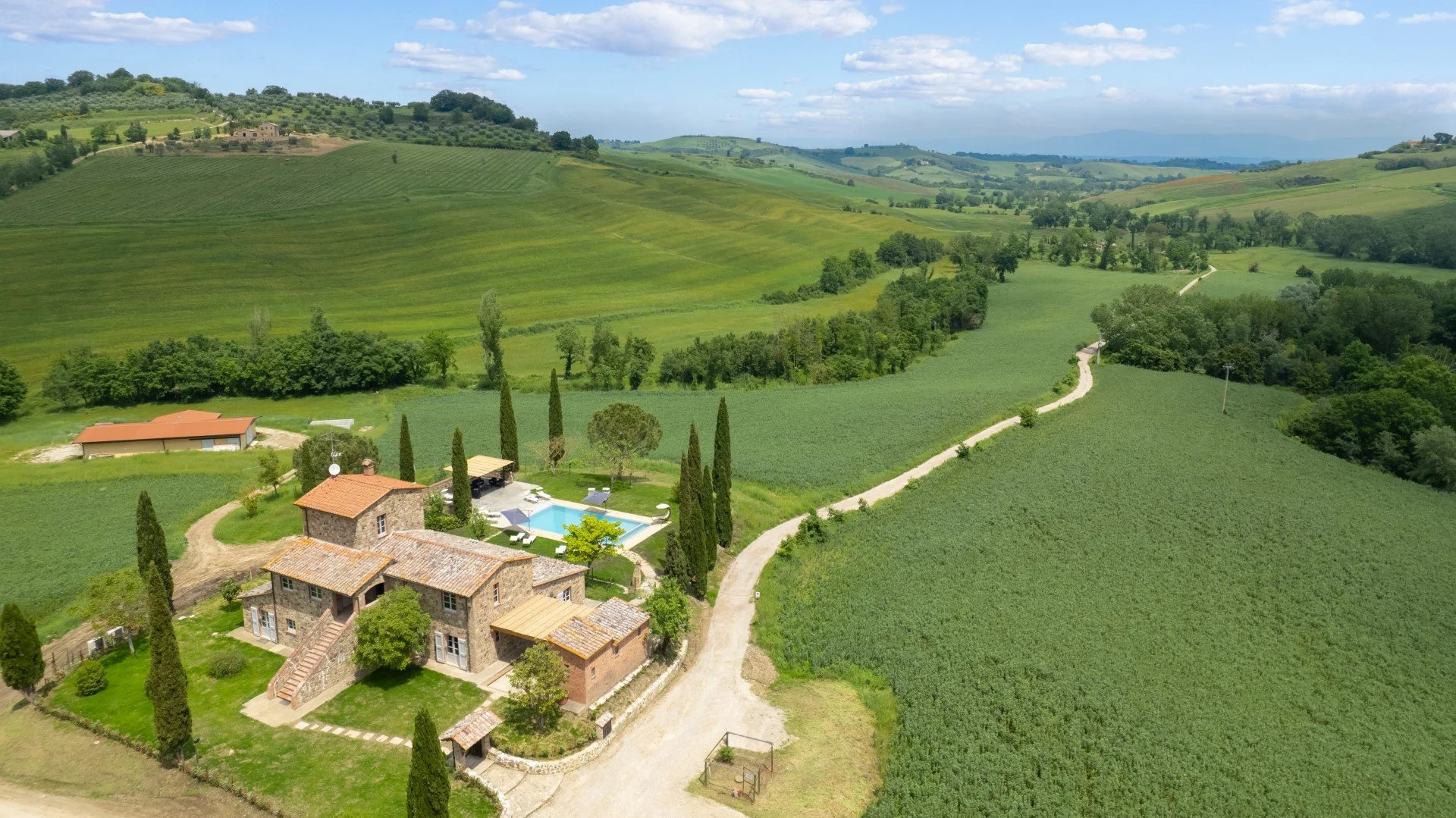 Aerial view of a countryside estate with stone house, swimming pool, cypress trees, and green rolling hills.